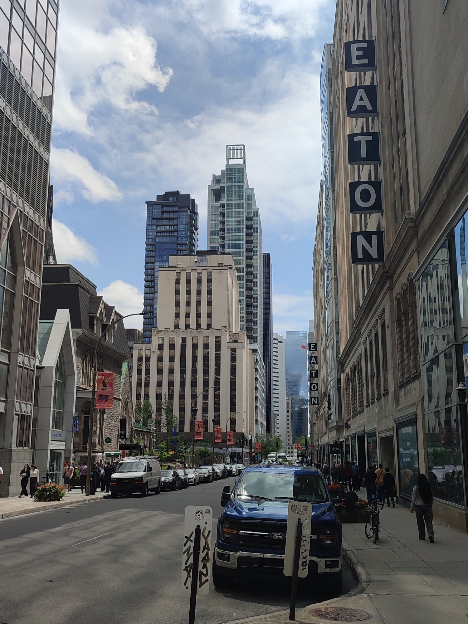 The image depicts a street scene in Montreal, Canada, on a bright, partly cloudy day. The view is down a city street lined with buildings of varying architectural styles. On the right side of the street, a large building with "EATON" signage vertically displayed is prominent. Its facade features a mix of stone and glass, with arched windows on the lower levels. The sidewalk in front of this building is populated with pedestrians.
On the left side of the street, modern and older buildings coexist. One building has a glass facade with a grid pattern. Further down, a more ornate, older structure with arched windows and a pointed roof is visible. Several flags or banners hang from poles along the sidewalk.
The street itself is paved and appears to be a one-way road with cars parked along both sides and traffic moving down the center. Several vehicles are visible, including a blue Ford pickup truck in the foreground and a white van further up the street. Pedestrians are walking on the sidewalks and crossing the street. The sky is blue with scattered white clouds.