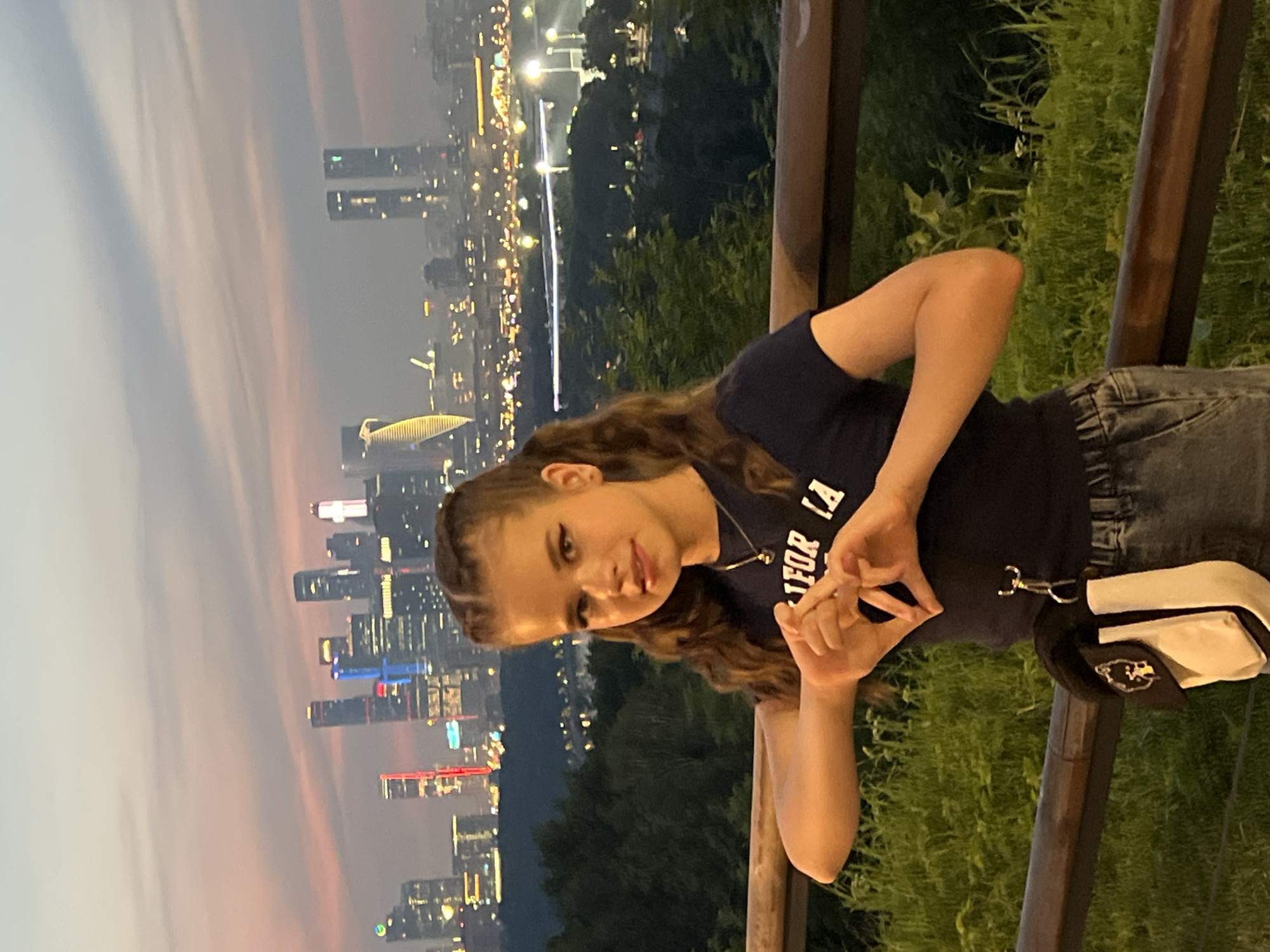A young woman with long, wavy brown hair styled in two braids stands in front of a cityscape at dusk. She is wearing a dark t-shirt with a graphic print, jeans, and a small crossbody bag. Her hands are clasped together in front of her chest, forming a heart shape. The background features a panoramic view of Moscow, Russia, with illuminated skyscrapers and city lights stretching into the distance. The sky is a soft blend of pink, orange, and gray, indicating either sunrise or sunset. The woman is positioned on a raised platform or overlook, with a wooden railing in the foreground. Lush green vegetation is visible around her, suggesting a park or elevated natural area within the city. The overall atmosphere is serene and picturesque, capturing a moment of personal expression against a vibrant urban backdrop.