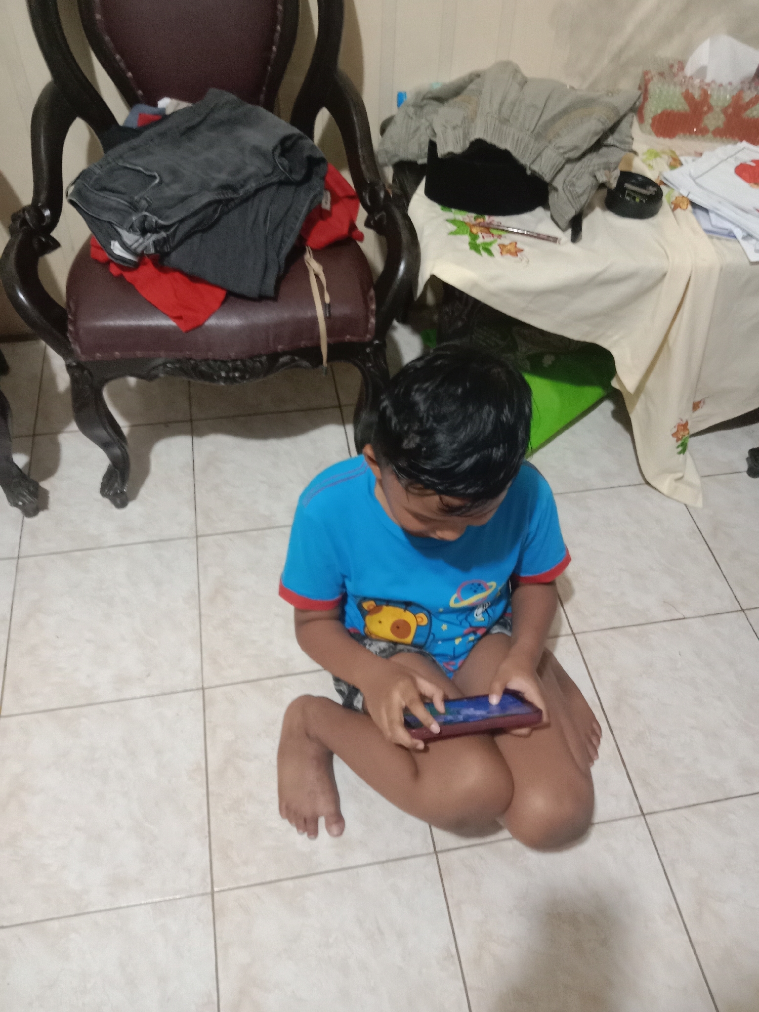 A young boy, possibly of Indonesian ethnicity, is sitting on a tiled floor, engrossed in playing a mobile game on his device. He is wearing a blue t-shirt with a graphic print, and his legs are crossed. The scene appears to be indoors, possibly in a home in Madiun, Indonesia. In the background, there is a dark wooden chair with some clothes piled on it, and a table covered with a light-colored cloth, upon which other items are placed. The lighting suggests an indoor setting, possibly during the day or evening with artificial light.