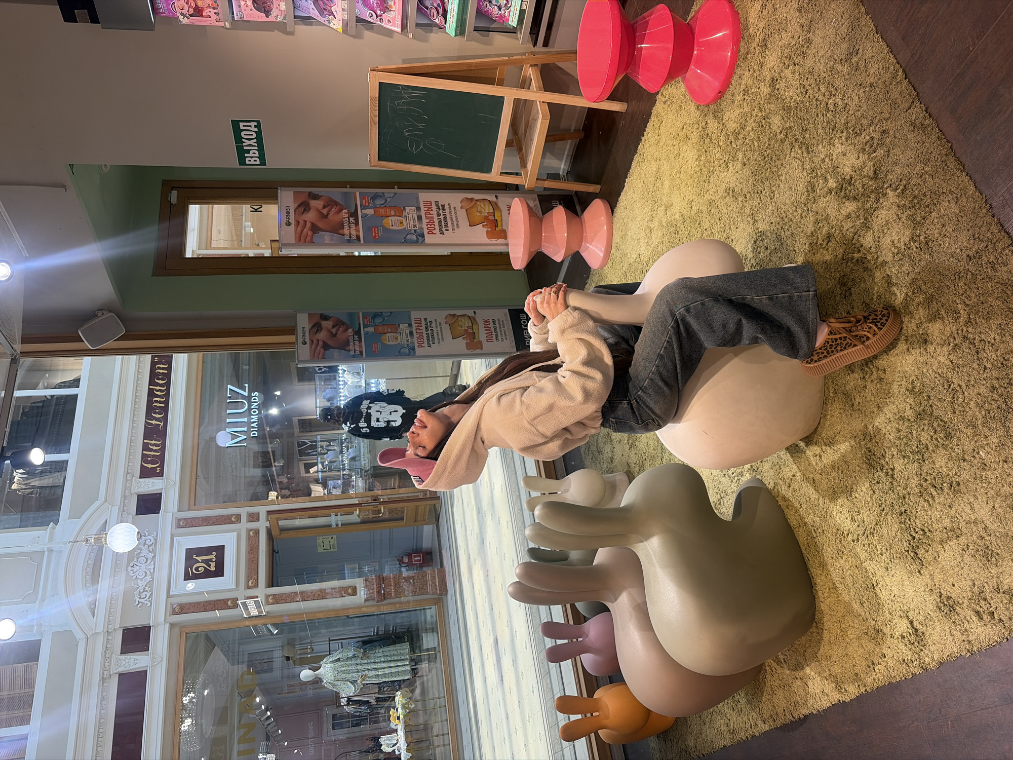 In Saint Petersburg, Russia, a young woman sits on a large, light-colored rabbit-shaped stool in a brightly lit indoor space. She is wearing a cream-colored hooded sweatshirt, blue jeans, and leopard-print platform shoes. Her pink baseball cap is pulled down over her head, and she is looking upwards with her mouth slightly open. The stool she is sitting on is part of a group of similar rabbit-shaped stools in various pastel colors, arranged on a textured green rug. Behind her, a chalk board stands on an easel, with some scribbled writing on it. To her right, there are two bright pink, hourglass-shaped stools. In the background, through large glass windows, a clothing store with mannequins and displays is visible, along with storefronts bearing Russian lettering, including "MIUZ DIAMONDS" and "Old London." A sign with the number "21" is also visible on one of the storefronts. The overall environment appears to be a commercial or retail setting, possibly a shopping mall or a boutique.