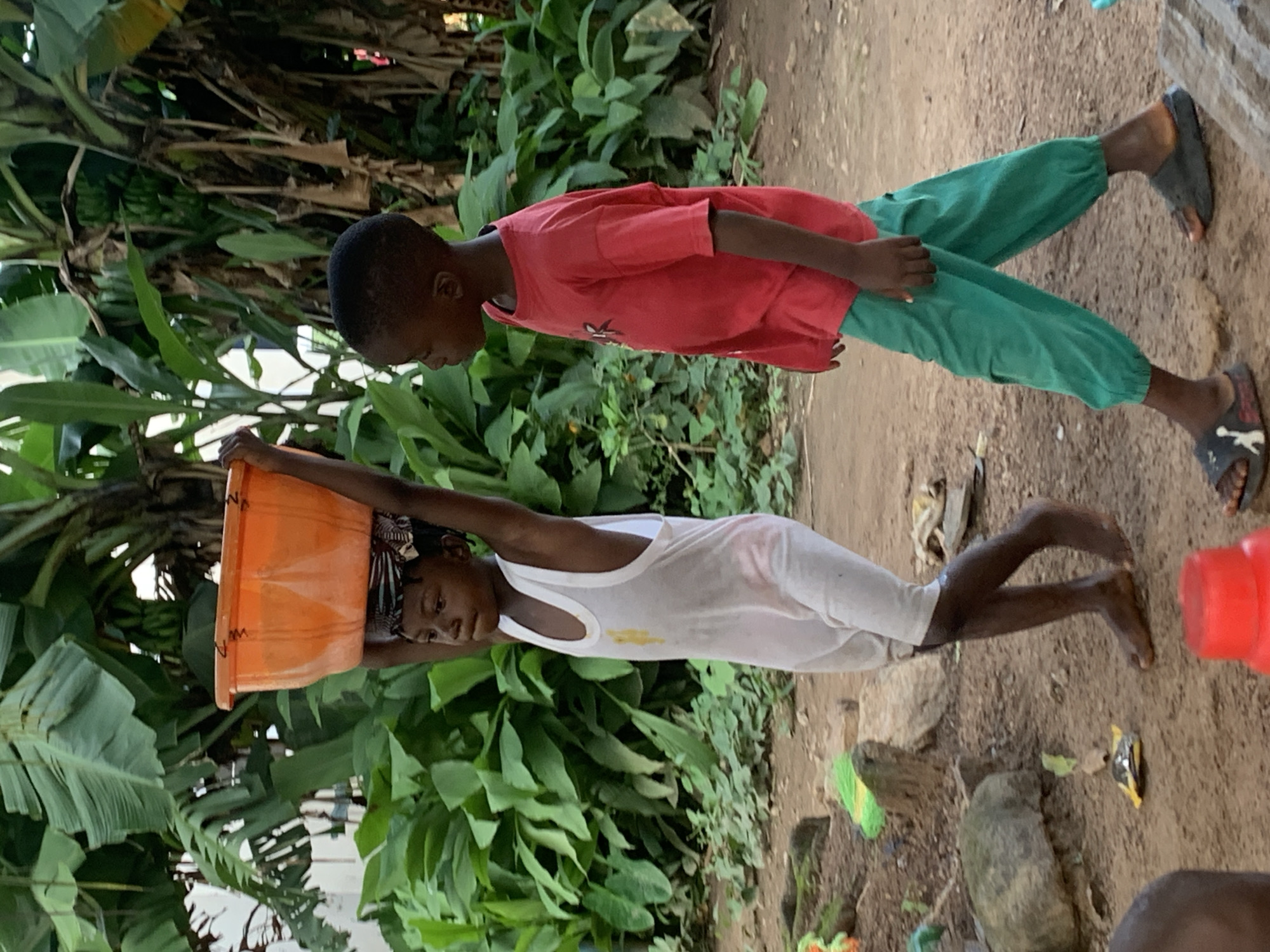 In a dusty, outdoor area with abundant green foliage and banana trees in the background, two young children are walking. The child on the left, wearing a white tank top and shorts, is balancing a large orange plastic basin filled with water on their head. A patterned cloth is wrapped around their head to help secure the basin. The child on the right, dressed in a red t-shirt and green pants, walks alongside. The ground is dirt, with scattered rocks and some discarded items like plastic wrappers. The overall scene suggests a daily chore or task in a rural or semi-rural environment within the city of Akwanga, Nigeria.