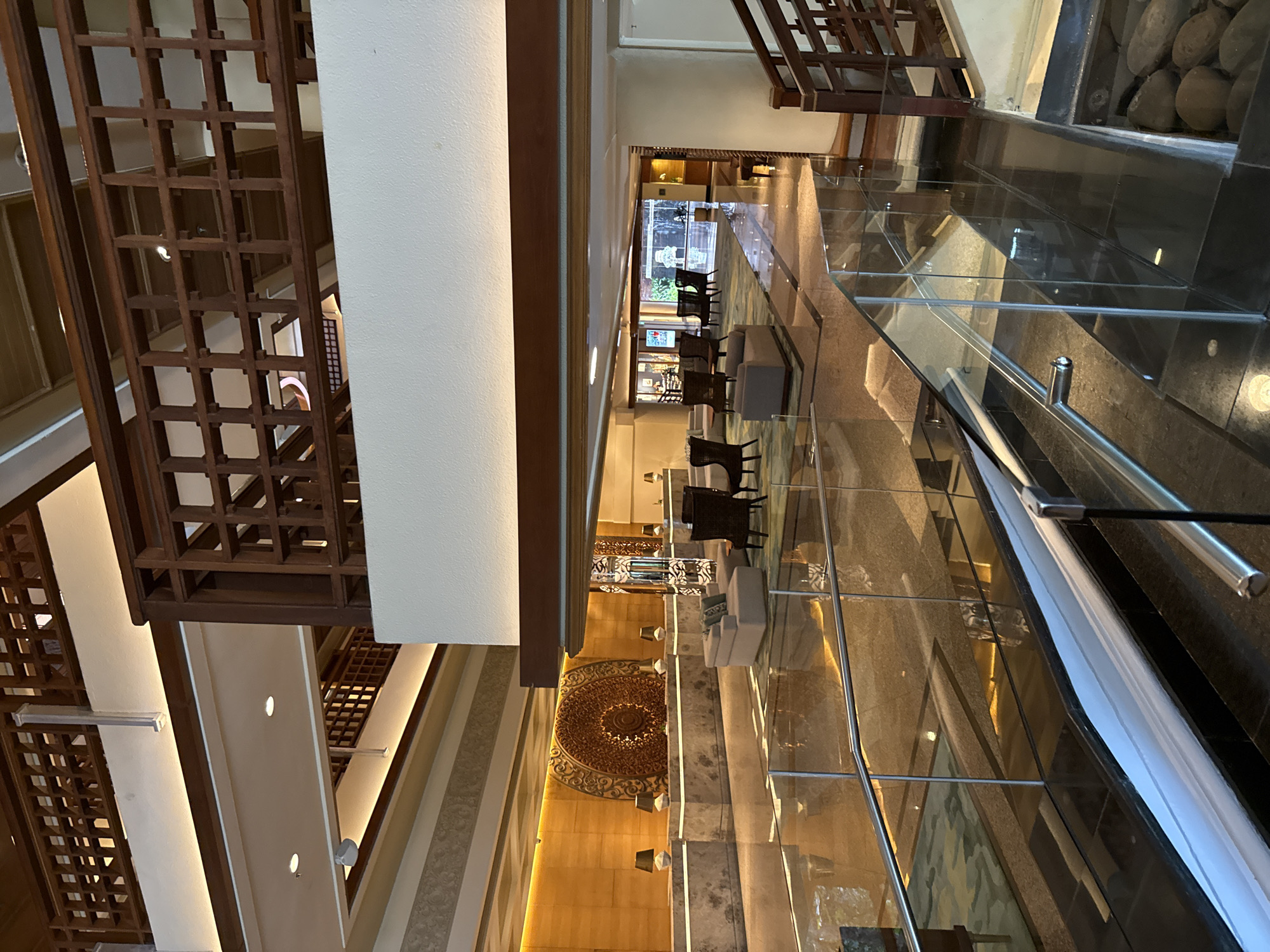This indoor shot shows the multi-story atrium of a building, most likely a luxury resort, in Nusa Dua, Indonesia. The perspective is looking down into the atrium.
Architectural elements include a multi-story wooden lattice structure on the left, and a decorative wall with an ornate circular design on the lower right. A glass-floored walkway spans the width of the atrium, with a metal railing visible in the foreground on the right.
The lobby area contains seating arrangements with chairs and sofas. Further in the distance, the lobby leads to an entrance with a view of the exterior, visible through glass doors/windows.
The overall ambiance suggests an upscale environment. No individuals are visible in the frame.