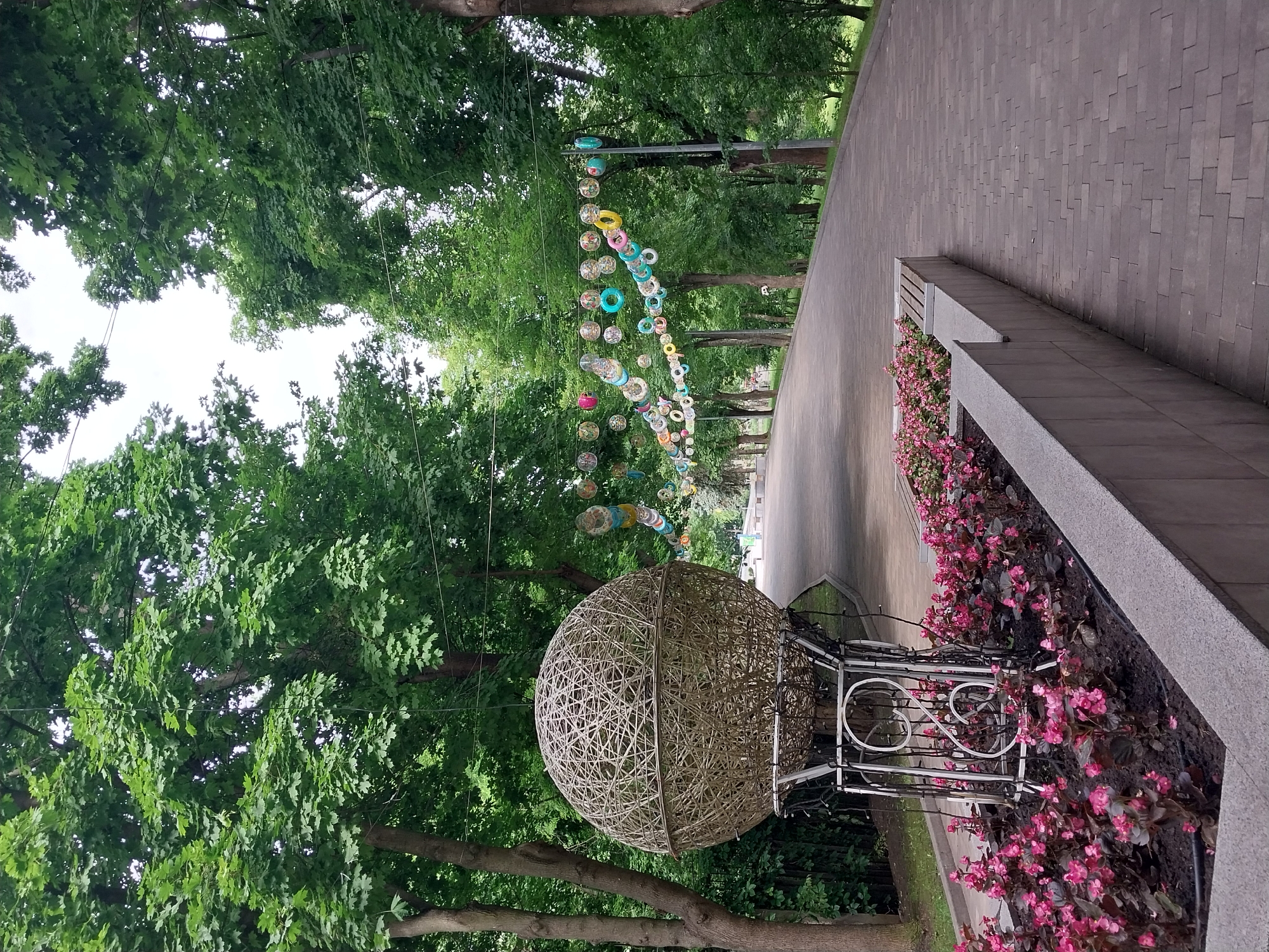 The image depicts a decorative pathway in a city park, likely Dnipro, Ukraine, featuring an artistic installation of a large woven sphere resembling a hot air balloon, adorned with lights. The pathway, paved with gray bricks, curves gently to the right, flanked by lush green trees and a border of vibrant pink flowers. Overhead, a string of colorful circular decorations, possibly rings, adds a whimsical touch to the scene. The overall ambiance suggests a well-maintained public space designed for leisure and aesthetic appeal. No people are visible in the frame.