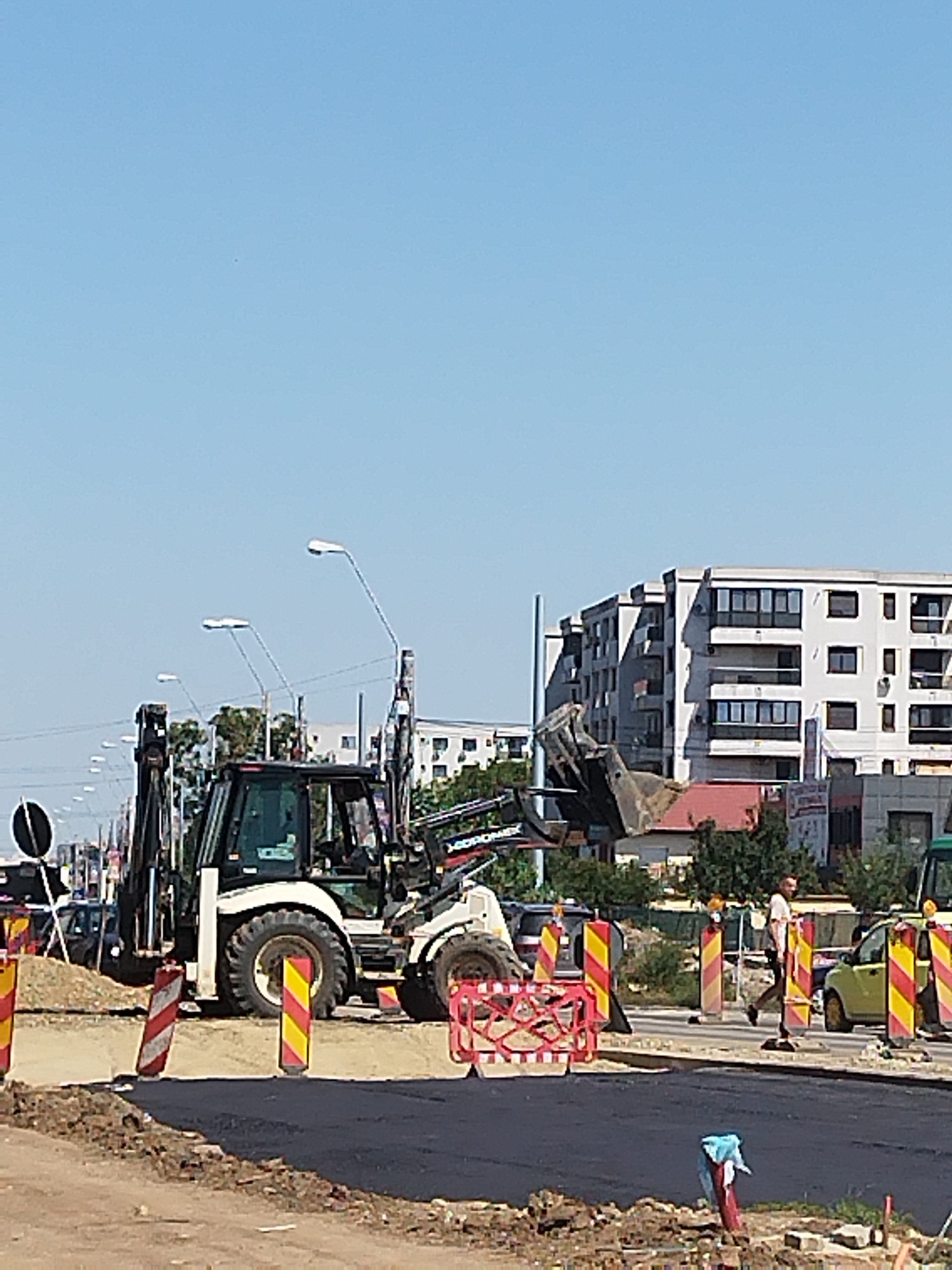 The image depicts a construction site on a clear day in Bucharest, Romania. In the foreground, freshly laid asphalt covers a section of road, bordered by loose soil and debris. Several traffic barriers, distinguished by alternating red and yellow stripes, are strategically placed around the work area. A white backhoe loader, a heavy construction machine with a scoop, is positioned on the site. A man in a white t-shirt and dark pants is seen walking across the street, and a yellow car is parked nearby. In the background, a multi-story apartment building with numerous windows and balconies stands against a bright blue sky. Several streetlights are visible along the road. The overall scene suggests ongoing road construction or repair work in a semi-urban environment.