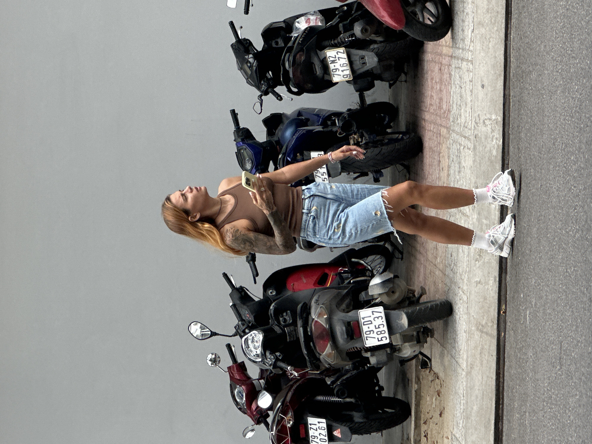 A woman stands on a sidewalk in Nha Trang Ward, Vietnam, holding a mobile phone. She is standing next to a row of parked motorbikes. The motorbikes have license plates with the registration code of "79", indicating they are registered in the Khanh Hoa Province.