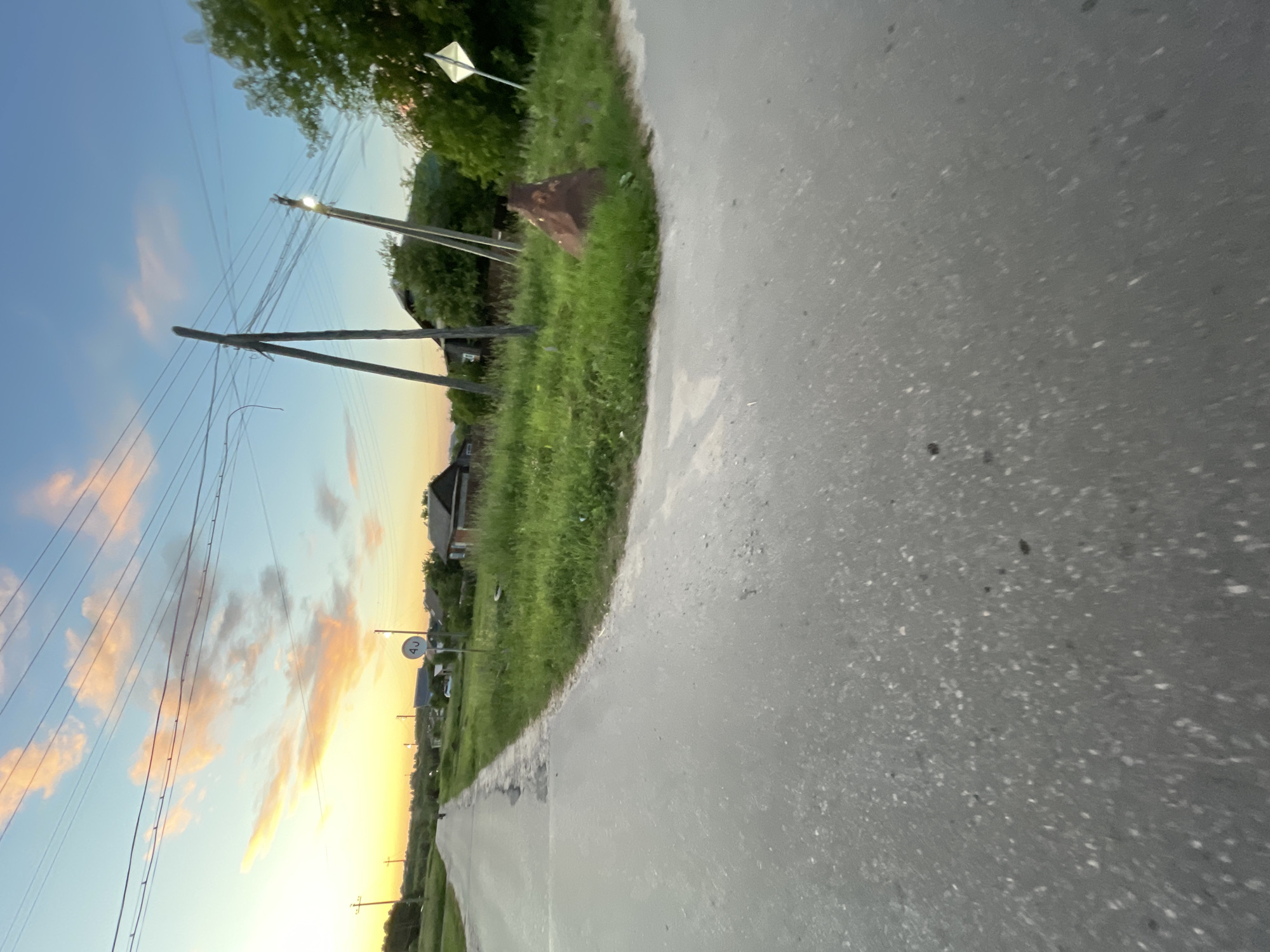 The image depicts a rural road in Krasnye Baki, Russia, at dusk. The road is paved and appears to be a two-lane road that curves to the left. On the right side of the road, there is a grassy verge with several wooden utility poles supporting power lines. One of the poles has a lit streetlamp. Further down the road, there are houses with dark roofs and some trees. A circular road sign with "40" inside it is visible on the right side of the road. In the distance, the sky is illuminated with warm hues of orange and pink from the setting sun, with scattered clouds. The overall impression is of a quiet, rural evening. There are no people clearly visible in the image.