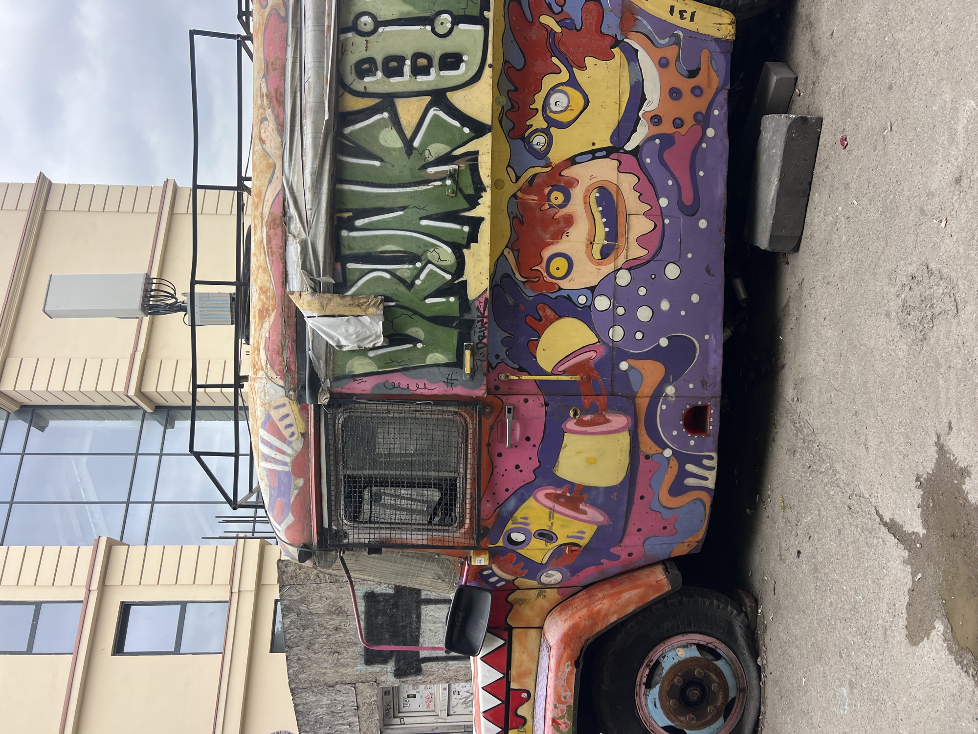 The image depicts the side of a brightly painted, graffiti-covered bus parked on a cracked asphalt surface. The bus is adorned with various cartoonish figures and psychedelic patterns in vibrant colors such as pink, purple, orange, and green. The word "DRUNK" is spray-painted in large green letters across the side. The front of the bus features a shark-like design. The front left tire is flat, and the bus is supported by two concrete blocks underneath its chassis. In the background, a modern building with large glass windows and beige facade stands, with an antenna structure visible on its roof. The sky is overcast. This scene is located in the city of Tbilisi, Georgia, specifically in the Vera district.