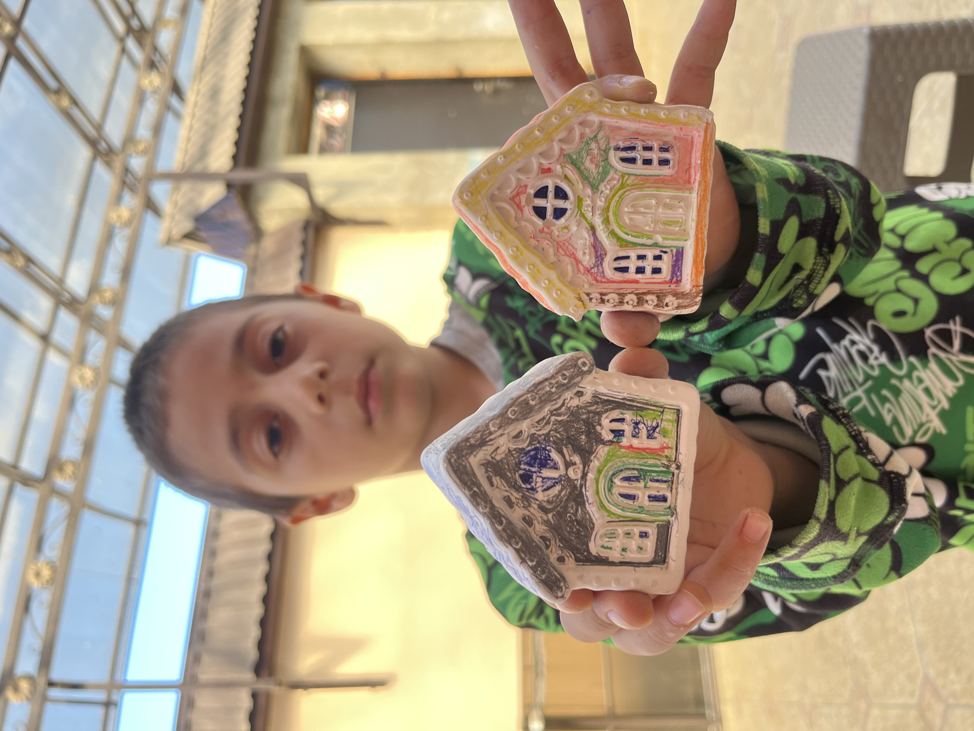 A young boy in Kaspiysk, Russia, is holding two painted clay house ornaments in his hands. The top ornament is decorated with pink, yellow, purple, and green paint. The bottom one is painted with black, gray, blue, and green. The boy is wearing a green and black patterned sweatshirt. A glass and metal structure is visible in the background. The scene appears to be outdoors.