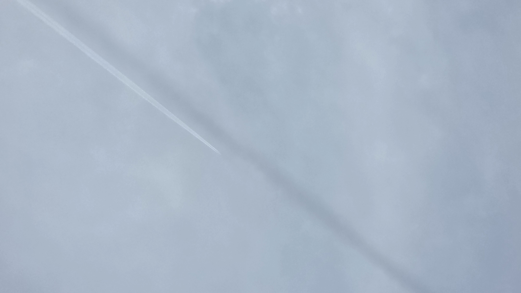 The image depicts a light blue sky with a faint airplane contrail visible in the upper left portion of the frame. There is also a long, hazy streak diagonally bisecting the image from the upper left to the lower right. The sky has a cloudy, diffused quality. The image was likely taken from Blagoevgrad, Bulgaria.
