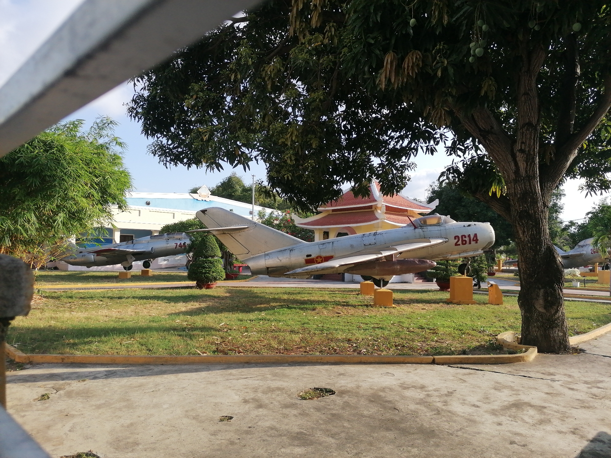 The image shows a park or memorial in Nha Trang Ward, Vietnam, featuring several retired MiG fighter jets. One jet, marked with the number "2614," is prominently displayed in the center, while others labeled "744" and "26" are visible in the background. The jets are placed on small concrete blocks on a grassy lawn. A large tree is present near the right side of the frame, partly obscuring the jet in the center. A traditional Vietnamese building is partially visible behind the jets. The foreground consists of a paved area with visible damage and repairs. A metallic fence is present on the left side of the image, partially obscuring the view. The scene appears to be taken in daylight with clear weather.