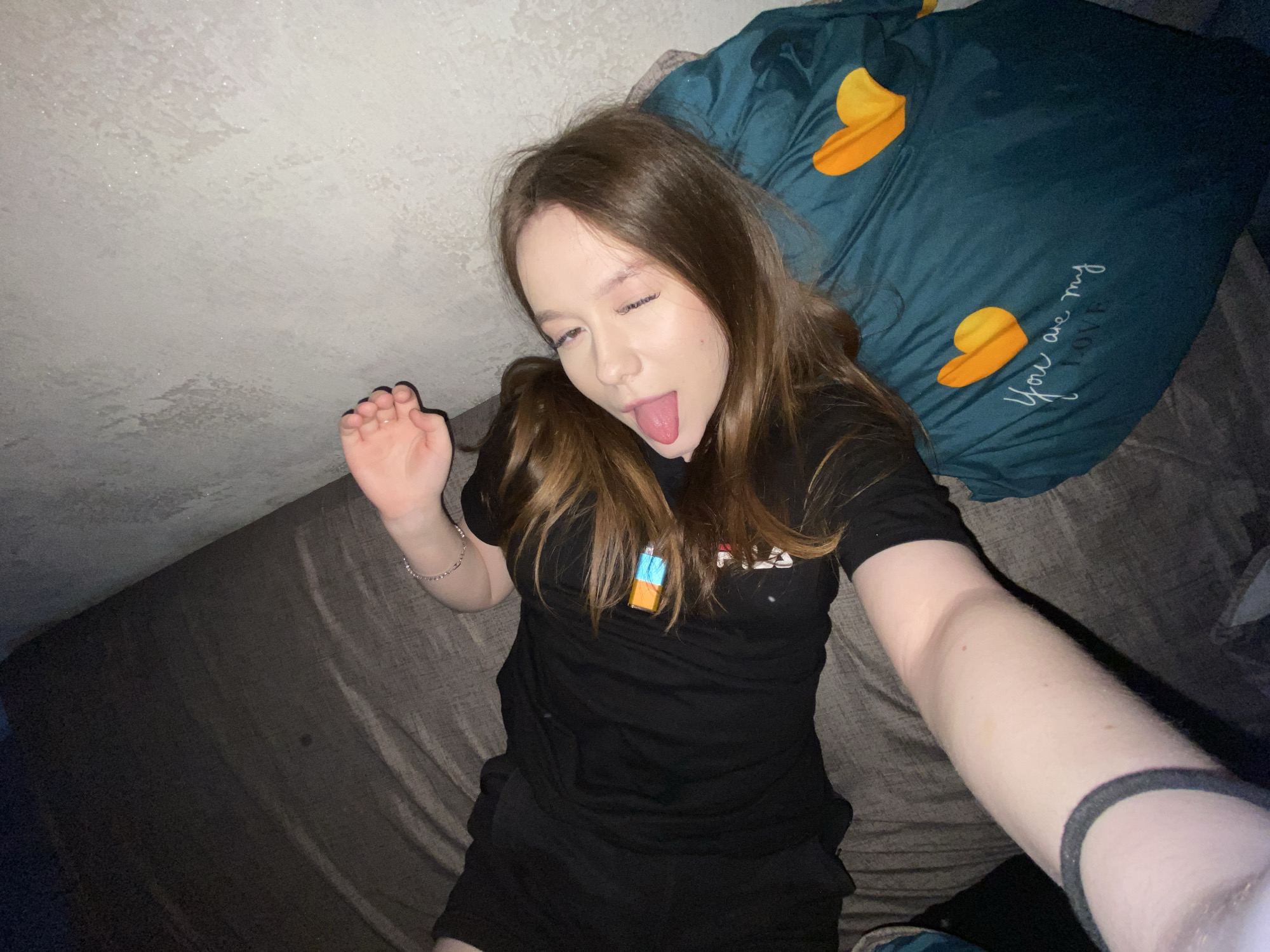 A young woman is lying on a bed with a dark brown sheet and a green pillow with yellow heart shapes and the words "You are my love" written on it. She is sticking out her tongue and winking. She has brown hair and is wearing a black t-shirt and black shorts. She has a bracelet on her left wrist. Her right arm is extended towards the camera. The background is a textured white wall. The location is Chaikovsky, Russia.