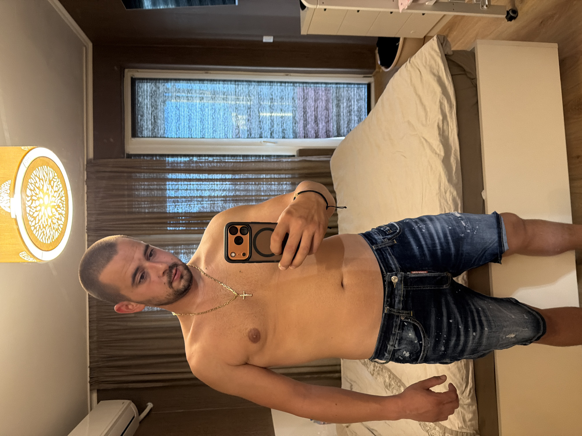 The media file shows a young man taking a selfie in a room. He is shirtless and wearing denim shorts. He has a gold chain with a cross pendant around his neck. The room appears to be a bedroom, with a bed and a window visible in the background. The window has blinds that are partially closed, and it is daytime. The man has a serious expression on his face. There is a lamp with a decorative shade on the left side of the image. The overall impression is that of a casual, personal moment captured by the man himself. The man's shorts have a label with the text "DSQUARED2".