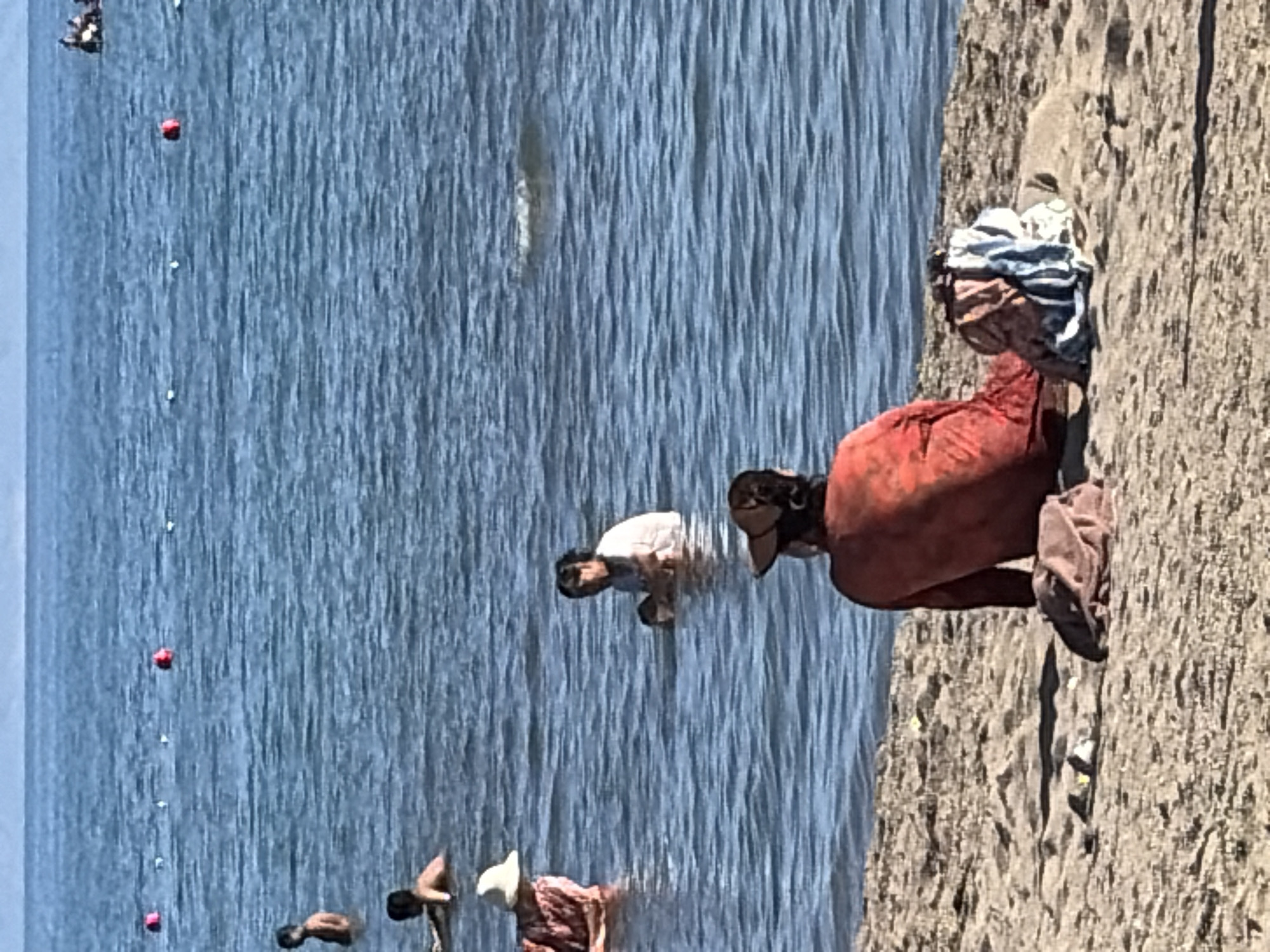 A medium shot shows a beach with people in the water and on the sand. In the foreground on the sand, a person wearing a red shirt and a straw hat sits facing away from the camera, with a pile of clothes next to them. In the middle ground, in the water, a person in a white shirt sits, and to their left, another person wearing a patterned shirt and a white hat sits. In the background, several people are in the water, and a red buoy is visible in the water. The sky is visible at the top of the image.