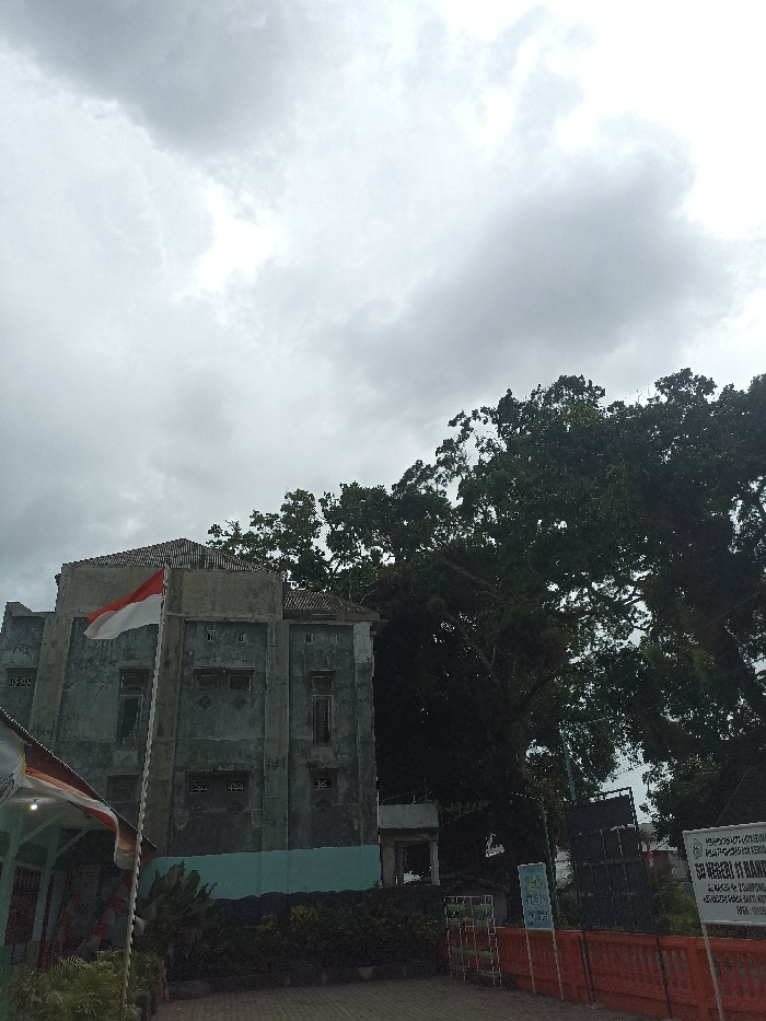 An outdoor scene in Lhokseumawe, Indonesia, captured under an overcast sky. The primary subject is a multi-story, weathered concrete building located left of center, featuring a red and white Indonesian flag on a pole attached to its facade. The building has multiple rectangular windows, some appearing closed or boarded, and a section of its lower exterior is painted light blue-green. To the right and behind the building, a dense canopy of dark green trees dominates the background. In the lower left foreground, a portion of a smaller structure with a white awning is visible. The ground appears paved. Along the mid-ground right, a series of orange-red barriers or fences are present, behind which stand multiple signs. A prominent white sign with black text identifies "SD NEGERI 11 BANDA" (State Primary School 11 Banda), along with additional smaller text below. Dark, box-like structures, possibly speakers, are visible behind the barriers. No individuals are depicted in the frame.