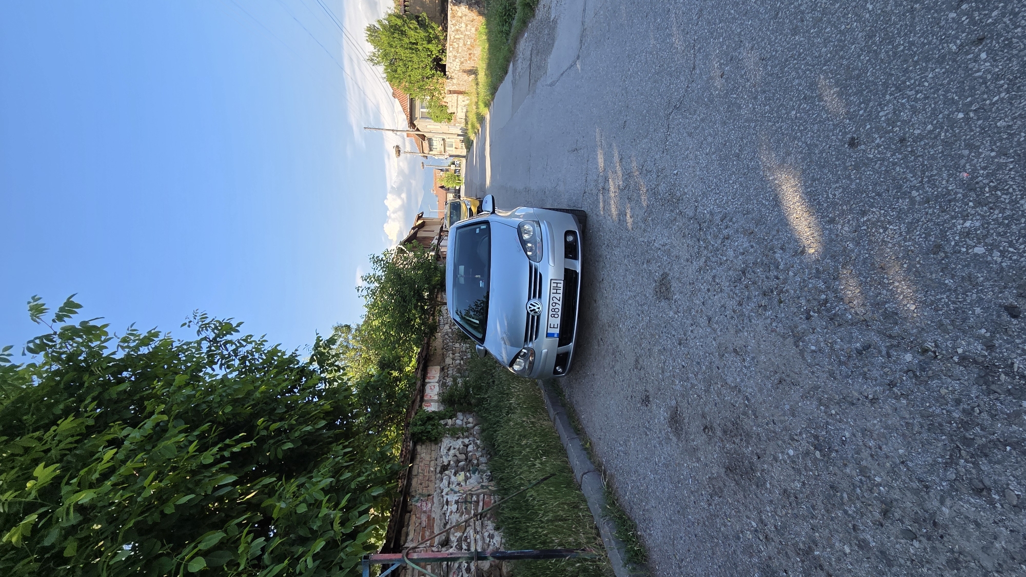 A silver Volkswagen Golf is parked on the right side of a narrow, paved road in a village. The car is positioned facing forward, with its front bumper and license plate, which reads "E 8892 HH", clearly visible. To the left of the car, a low, weathered stone wall runs along the edge of the road, topped with overgrown grass and greenery. A large tree with dense green foliage arches over the left side of the image, casting shadows on the road. In the background, more trees and traditional village houses with tiled roofs are visible under a clear blue sky. Another car, yellow in color, is parked further down the road. The overall scene depicts a quiet, rural setting.