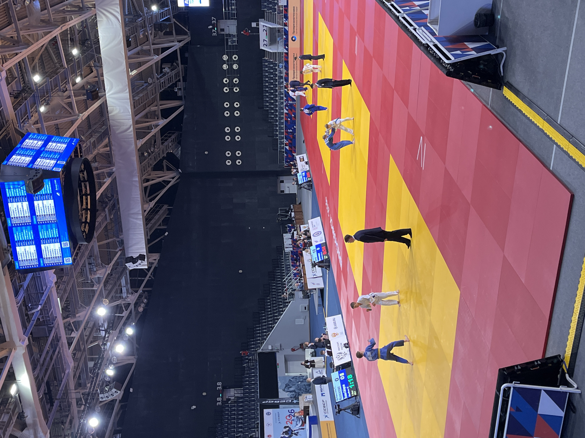 This image depicts an indoor judo competition held at the Platinum Arena in Krasnoyarsk, Russia. The scene is viewed from an elevated perspective, showcasing a vast arena floor covered with multiple red and yellow judo mats (tatami), surrounded by tiered seating that is largely unoccupied. The ceiling features exposed metal truss work and numerous bright overhead lights, illuminating the competition area.
In the foreground on the left, two young male judoka, one wearing a blue gi and the other a white gi, stand facing each other on a mat, observed by a male referee in a dark suit. Adjacent to this mat is a digital scoreboard displaying "Добрынин С." with a score of 0, "Новосельцев О." with a score of 1, and a remaining time of "1:18".
Towards the center and right, at least three other judo matches are actively in progress, with pairs of judoka (one in blue, one in white gi) engaged in grappling, each supervised by a referee in a dark suit. Additional mats with people are visible further in the background.
A large, multi-sided LED scoreboard is suspended from the arena ceiling, displaying multiple match results, competitor names, and scores in Russian. Various sponsorship and official logos are visible around the competition area, including "СЕВЕР" (Sever), "МИНСПОРТ РОССИИ" (Ministry of Sport of Russia), "JU.DO.RU", and "Промрезинотехника" (Promrezinotekhnika). The arena walls are dark, marked with section numbers such as "3.8 - 3.8" and "3.7 - 3.7". Yellow and black cable protectors run along the floor at the perimeter of the mats. The overall environment is set up for a professional sports event.
