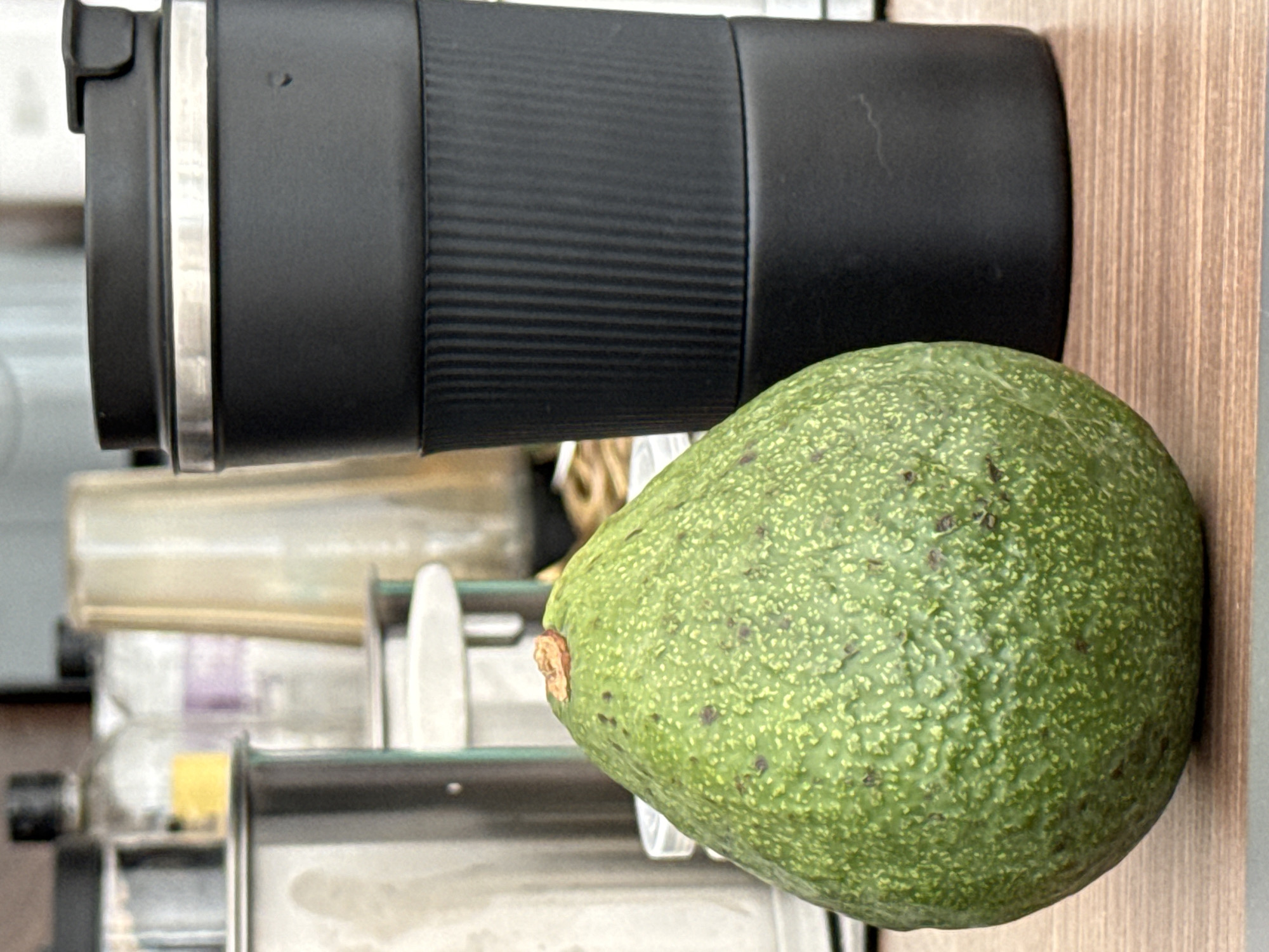 This media file captures a close-up of a whole avocado placed next to a black reusable travel mug. The avocado is vibrant green with a bumpy texture and some brown speckling, indicating its ripeness. The travel mug is cylindrical and appears to be made of matte black material with a ribbed section for grip. A silver band encircles the top of the mug.
In the background, out of focus, are hints of a kitchen or cafe setting. There are glimpses of stainless steel appliances, possibly a coffee machine or blender, and some glass bottles or containers. The lighting suggests it's daytime, possibly indoors with artificial light or near a window. The overall scene is simple and focused on the food and drink items. There is no visible text in the image. The image has been rotated 90 degrees clockwise.