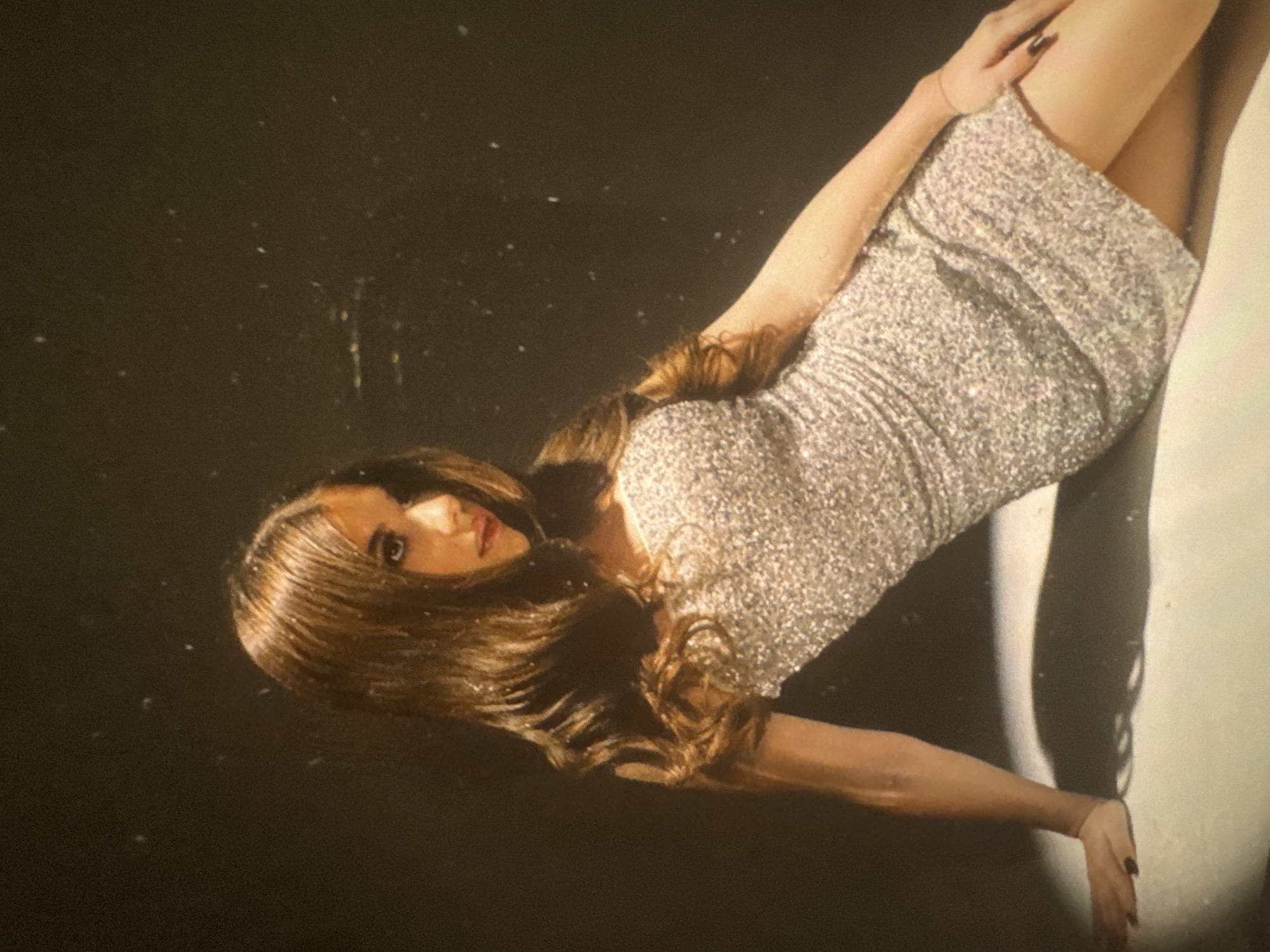 The image features a woman posing, captured from an overhead perspective and rotated diagonally, suggesting she is lying or leaning on a surface. She has long, wavy, rich brown hair that cascades around her head and shoulders. Her head is positioned towards the upper left of the frame, and she looks directly at the viewer with a calm and composed expression.
She is wearing a short-sleeved mini-dress that shimmers brightly, appearing to be covered in silver or light-gold sequins or glitter. Her arms are positioned with one hand resting on her upper thigh, and her legs extend towards the right.
The background on the left side is a dark, plain surface, likely black or deep brown, which is covered with numerous small white specks resembling dust or glitter. On the right side of the image, behind her lower body, there is a starkly contrasting vertical strip of a lighter, possibly white or off-white, surface. A distinct, elongated shadow of the woman's body is cast upon this lighter background, suggesting strong directional lighting from the right. There is no visible text in the image.