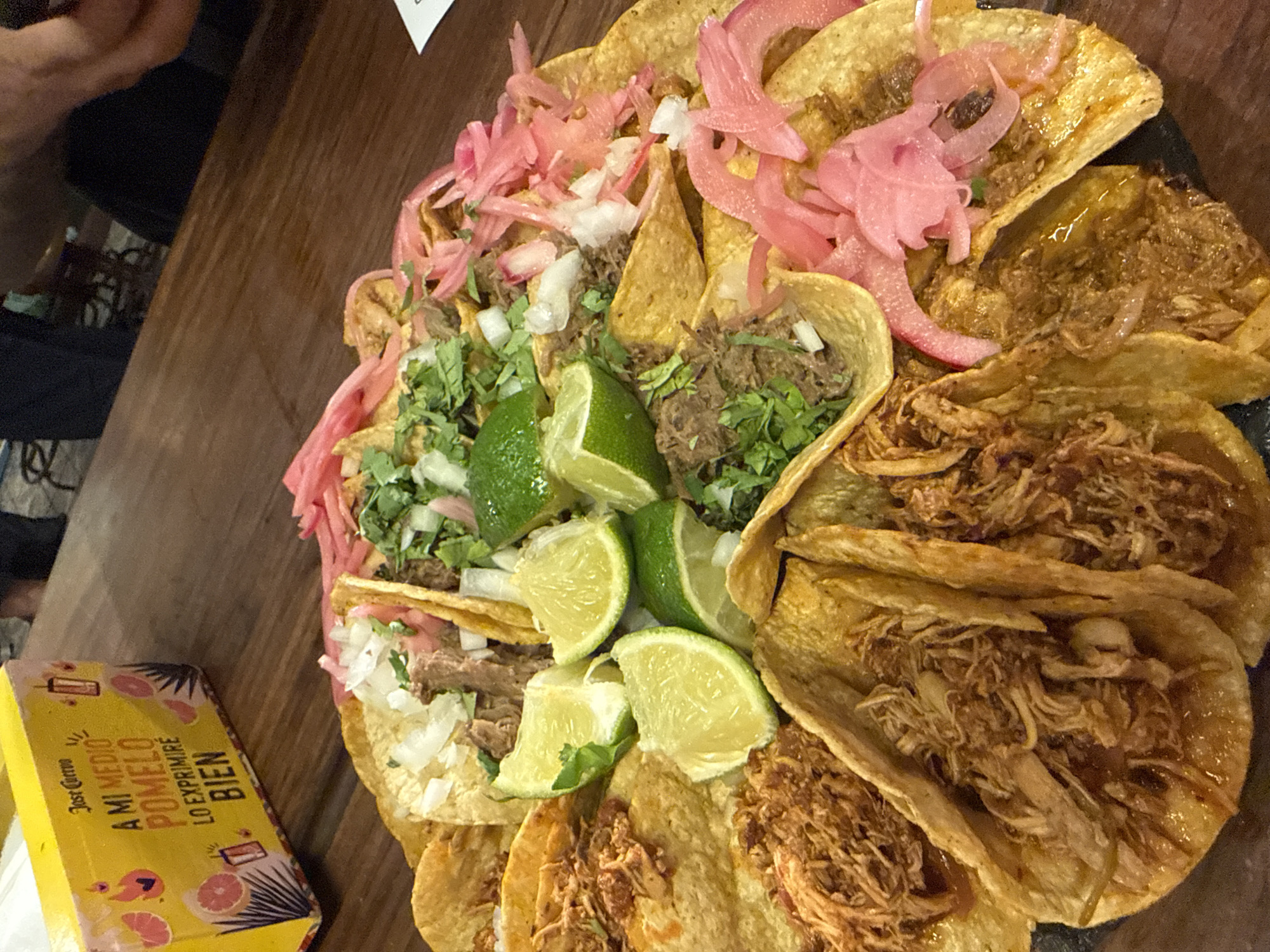 A platter of tacos is presented on a dark surface, likely a table, with a wooden texture visible in the background. The tacos are arranged in a circular fashion and appear to be filled with shredded meat, possibly pork or beef, and topped with chopped onions, cilantro, and pickled red onions. Lime wedges are scattered among the tacos. Some of the tortillas have a crispy, golden-brown appearance. A yellow box with Spanish text and graphics is partially visible in the lower left corner. The scene suggests a meal, likely lunch or dinner, in a casual dining setting. The lighting appears to be indoor, possibly from overhead lights or natural light from a window, casting a warm glow on the food. There is a hint of a person's arm and shoulder in the upper left corner, suggesting they are at the table or serving the food. The text on the yellow box reads "A MI MEDIO POMELO LO EXPRIMIRÉ BIEN", which translates to "My half grapefruit, I'll squeeze it well". It also features the brand "Jose Cuervo". The overall impression is one of a delicious and inviting culinary presentation.