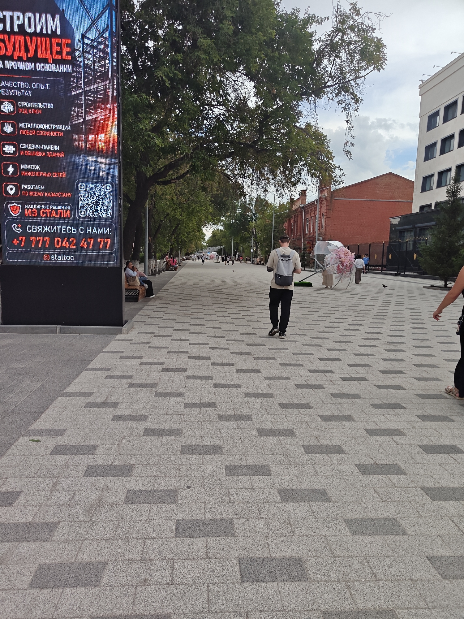The image depicts a wide, paved pedestrian walkway in a city, likely Petropavl, Kazakhstan. The walkway is paved with light grey and darker grey rectangular tiles arranged in a repeating pattern. On the left side of the walkway, a large, illuminated digital billboard displays advertisements and text in Russian, including the company name "СТРОИМ БУДУЩЕЕ" (Building the Future). The billboard also lists various services and contact information, including a phone number starting with "+7 777" and a social media handle "@staitoo".
Several people are visible on the walkway. In the foreground, a person wearing a light-colored t-shirt and dark pants, with a backpack, walks away from the camera. Further down the walkway, a person is seen pushing a large, clear umbrella decorated with pink artificial flowers. Other pedestrians are scattered along the walkway, some sitting on benches under trees.
The environment includes mature trees with green foliage lining the walkway, providing shade. In the background, brick buildings and a modern white building with multiple windows are visible. The sky is cloudy. The overall impression is that of a public urban space on a daytime.