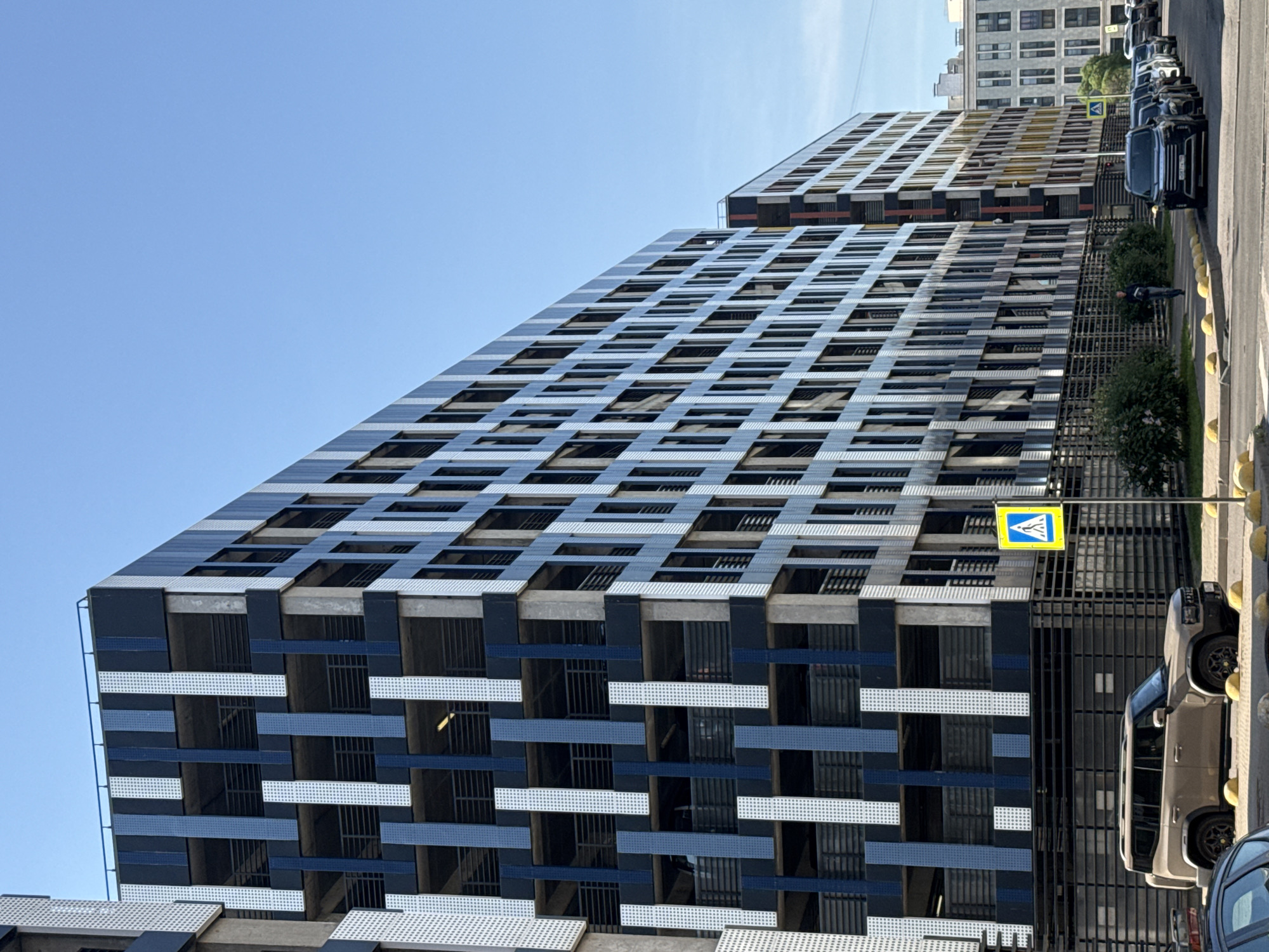 An eye-level shot captures a modern, multi-story building with a distinctive facade composed of alternating dark blue and white vertical panels interspersed with grey concrete structural elements. The building, likely a parking garage due to its open-air levels and visible railings, is situated in an urban environment. In the foreground, a light brown SUV is parked parallel to the street, facing left. The street itself is paved, with yellow bollards lining the curb. A pedestrian crossing sign with a yellow background and a white pedestrian symbol is visible to the right of the SUV. Beyond the main building, another similar structure extends further down the street, and further still, a residential building with a lighter facade and numerous windows can be seen. The sky is a clear, bright blue, suggesting fair weather.