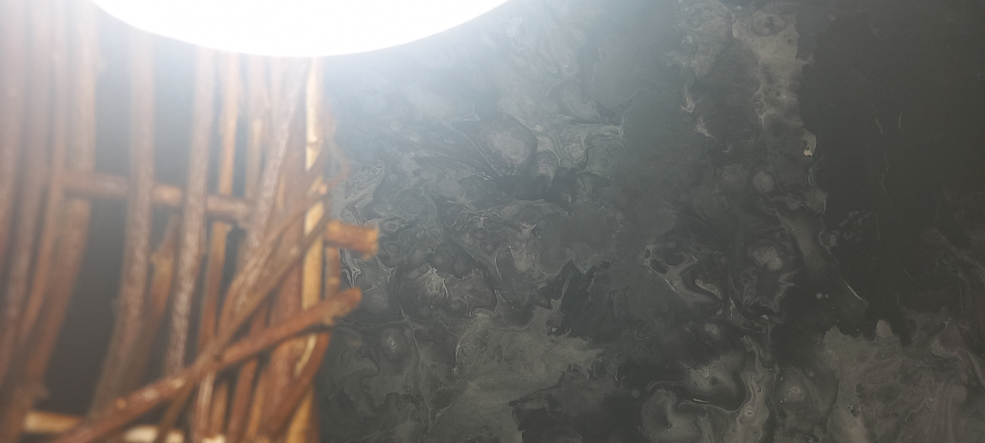 The image depicts a close-up, high-angle view of two distinct textures under bright illumination. The upper portion of the frame, approximately one-quarter of the total height, shows a blurred arrangement of natural, reddish-brown wooden elements, appearing as interwoven or horizontally stacked branches or twigs, consistent with a wicker structure or a natural, rough-hewn surface. These elements are out of focus. Below this, occupying the majority of the frame, is a dark, abstract pattern resembling a fluid art medium, such as an acrylic pour or marbled surface. This pattern features intricate swirls, organic cells, and interconnected shapes in varying shades of dark gray, black, and white. A powerful, undefined light source is prominently visible on the right side of the image, casting a significant, bright white glare across the scene, particularly affecting the mid-right section of the abstract pattern. No people, discernible objects other than the described textures, or specific actions are present. The visual content of the image does not contain specific landmarks, signage, or contextual elements to ascertain its precise geographical location within Kryvyi Rih, Ukraine.
