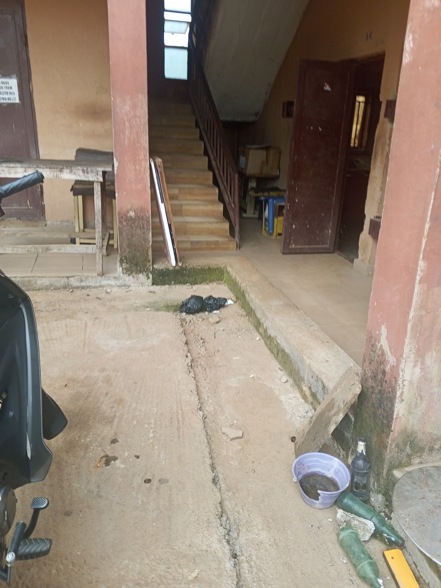 The image depicts an outdoor scene in what appears to be an urban environment, possibly in Abuja, Nigeria. In the foreground, a portion of a black motorcycle is visible on the left. The ground is covered in concrete, with some dirt and debris scattered about. To the right of the motorcycle, a black trash bag lies on the ground. Further to the right, a broken purple plastic bucket contains some dark liquid, next to which are three glass bottles, two green and one dark, lying on their sides. There are also some yellow and silver metallic objects on the ground.
In the background, a staircase with wooden railings leads upwards. The walls of the building are painted in earthy tones, with some areas showing signs of wear and tear, including moss or algae growth near the base. A wooden shelf unit is positioned against one of the walls. Further back, a dark brown door is visible, slightly ajar, revealing a glimpse of an interior space. Above the staircase, a window with multiple panes is open to the sky. A sign with text is affixed to a door on the far left of the image, but the text is illegible. The overall impression is of a utilitarian, somewhat neglected outdoor area adjacent to a building.