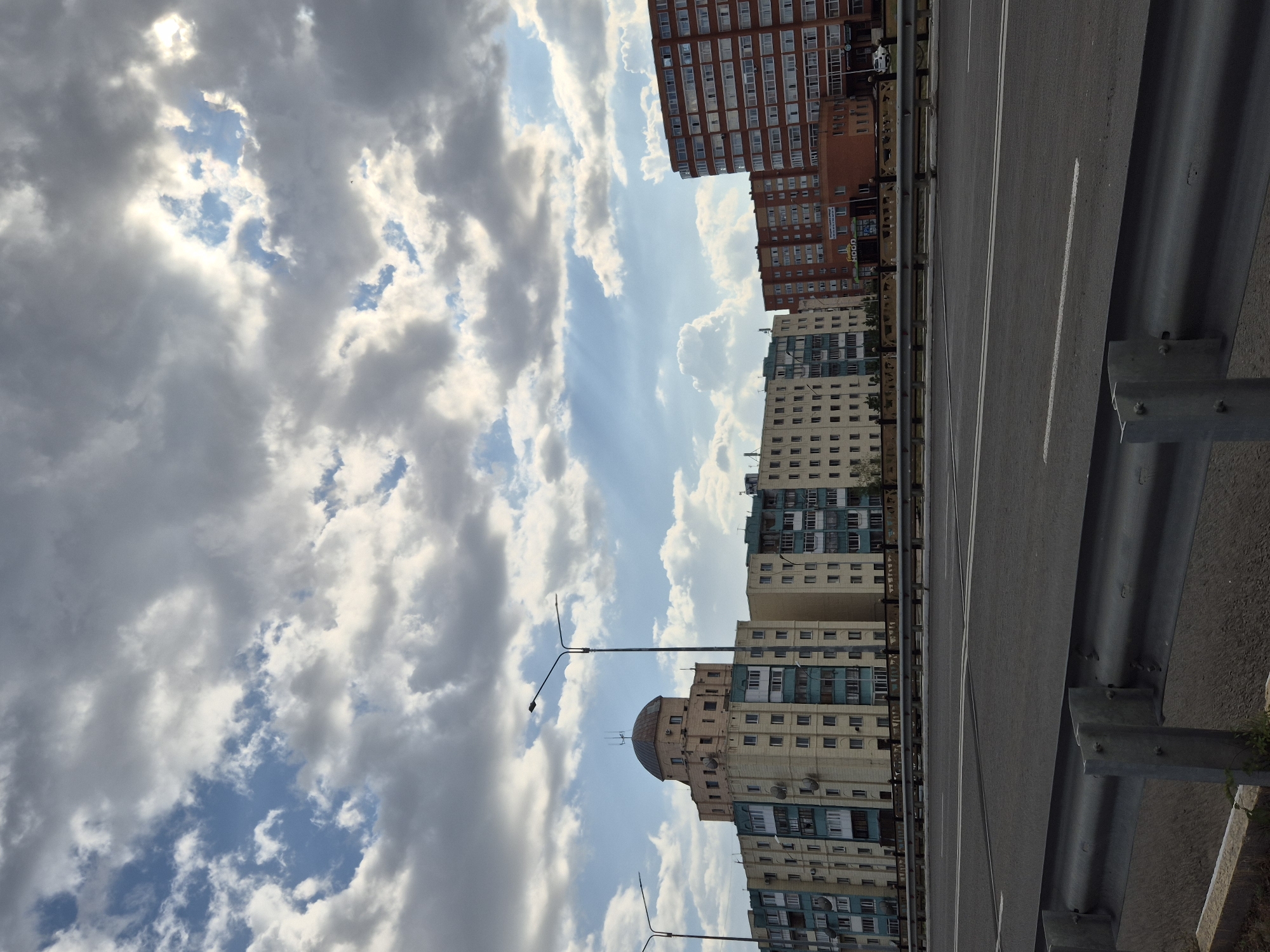 The image shows an urban scene with a multi-lane road in the foreground and a line of apartment buildings in the background. The sky is filled with dramatic clouds, with patches of blue sky and sunlight breaking through. The apartment buildings are of varying architectural styles, with one prominent building in the center featuring a domed roof. Streetlights are visible along the road. The scene appears to be captured from an elevated position, possibly from a bridge or overpass. Based on architectural styles and urban layout, this scene is likely located in Kokshetau, Kazakhstan.