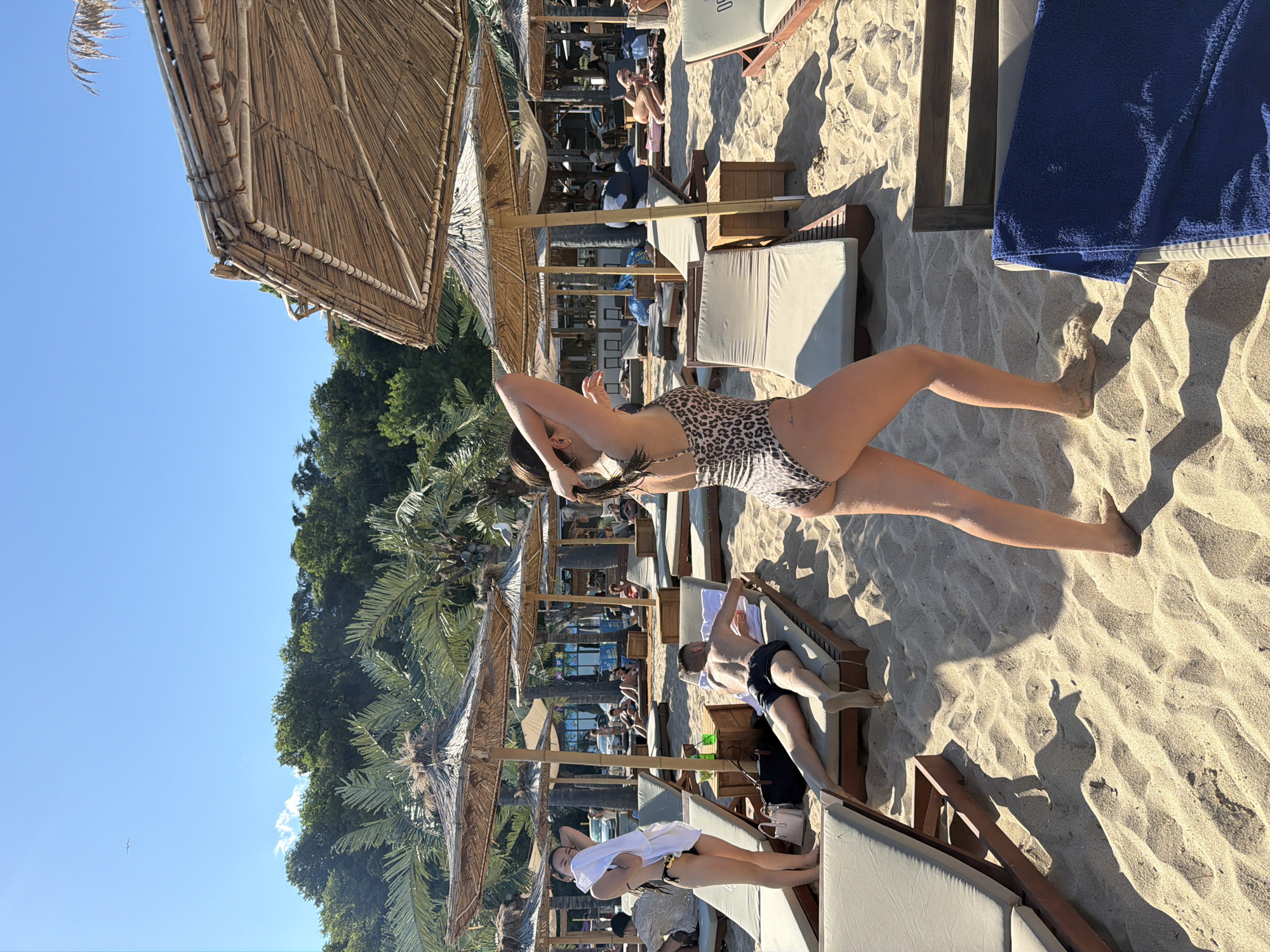 This image captures a scene at a beach resort in Varna, Bulgaria, under a clear blue sky. Several straw-covered umbrellas provide shade over lounge chairs on the sandy beach. In the foreground, a woman in a leopard-print swimsuit is standing, facing away from the camera with her arms raised and hands near her head. To her left, a man in black swim trunks lies on a lounge chair, his back to the camera. Another woman in a black bikini and a white towel is also visible to the left, standing near a lounge chair. In the background, more lounge chairs, umbrellas, and a line of trees and palm trees are present, with a glimpse of buildings or structures further back. Some people are seen relaxing on lounge chairs in the mid-ground and background, and a car is visible in the distance. The overall atmosphere is one of a sunny, recreational day at the beach.