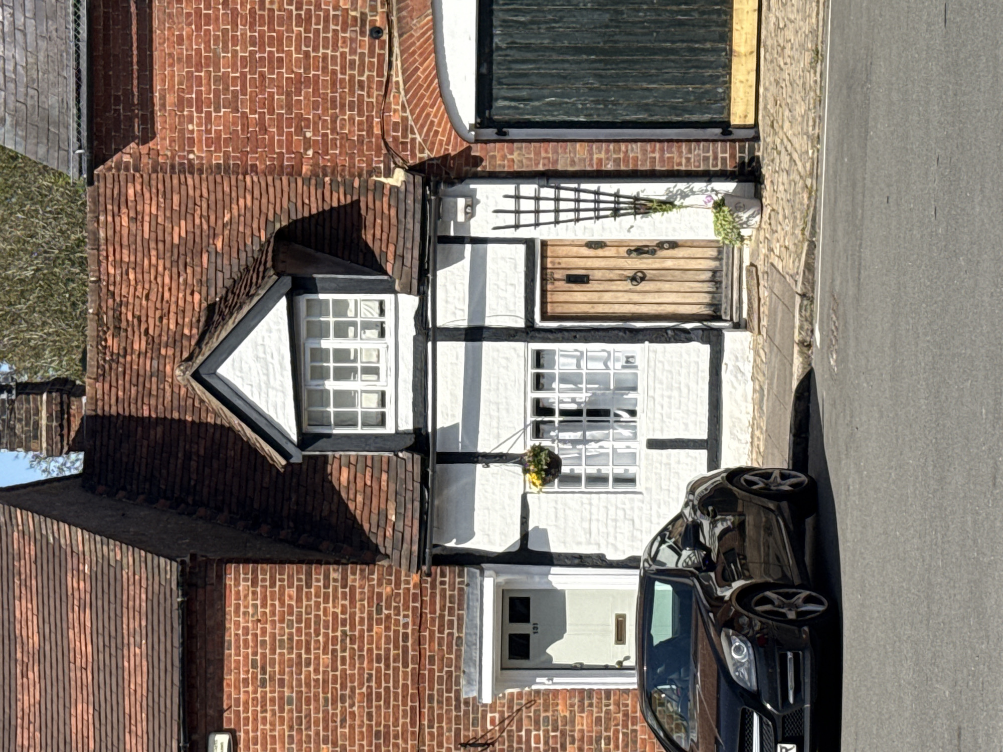 The image depicts a street scene in Amersham, United Kingdom. A two-story building with a half-timbered facade, characterized by white walls with black timber framing, is prominently featured. The building has multiple windows with white frames, a wooden door, and a brick roof. A black car is parked adjacent to the building on the asphalt road. The environment appears to be a residential area.
