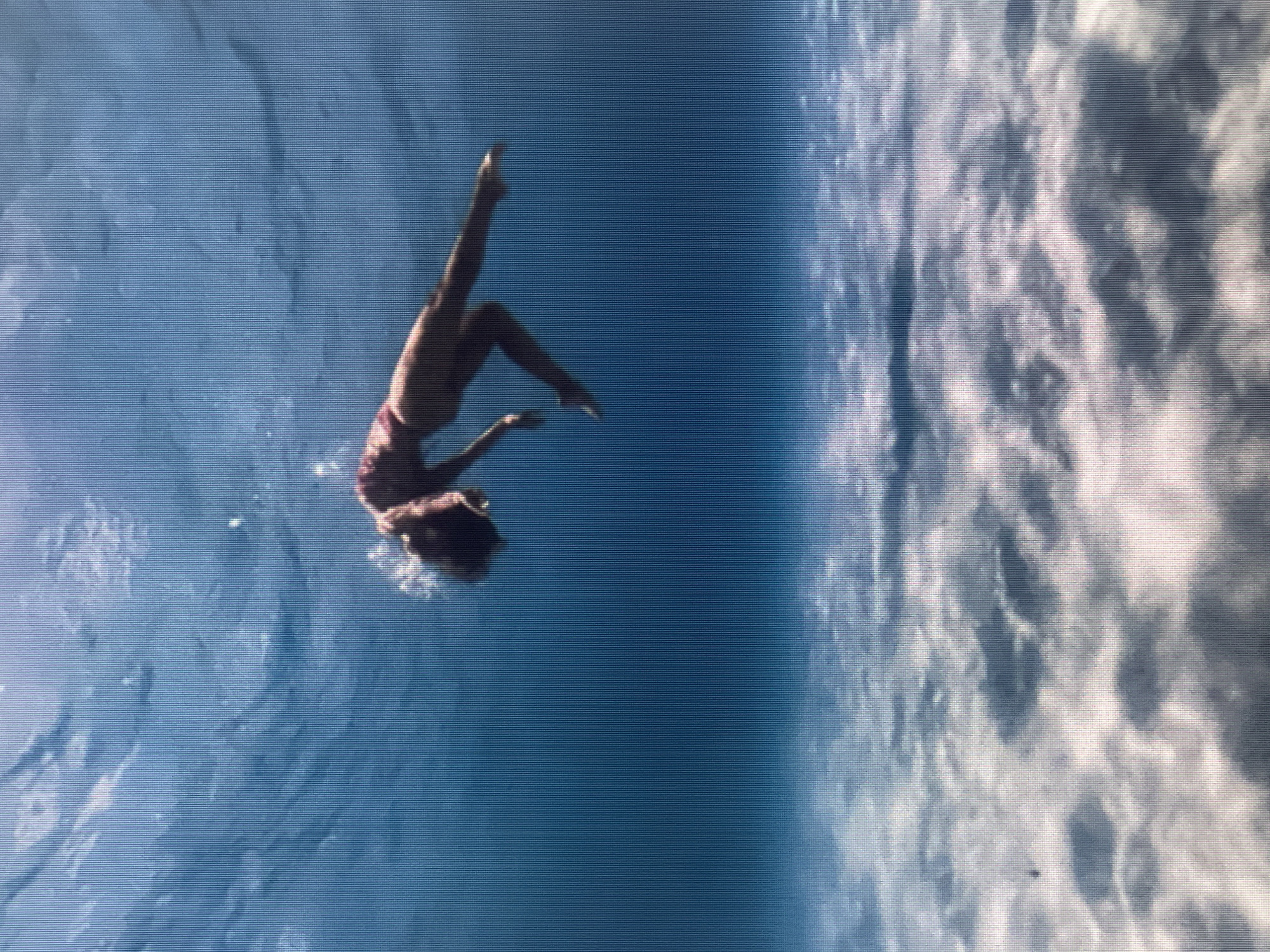 An underwater shot depicts a person floating gracefully in clear blue water, with sunlight filtering down from above, illuminating the sandy seabed below. The person, wearing a pink swimsuit, is suspended horizontally in the water, with their body arched backward and arms extended. Their head is tilted back, and their hair fans out around them, creating a sense of peaceful surrender to the water's embrace. The scene evokes a feeling of tranquility and freedom, as the person appears to be weightless and at one with their aquatic environment.