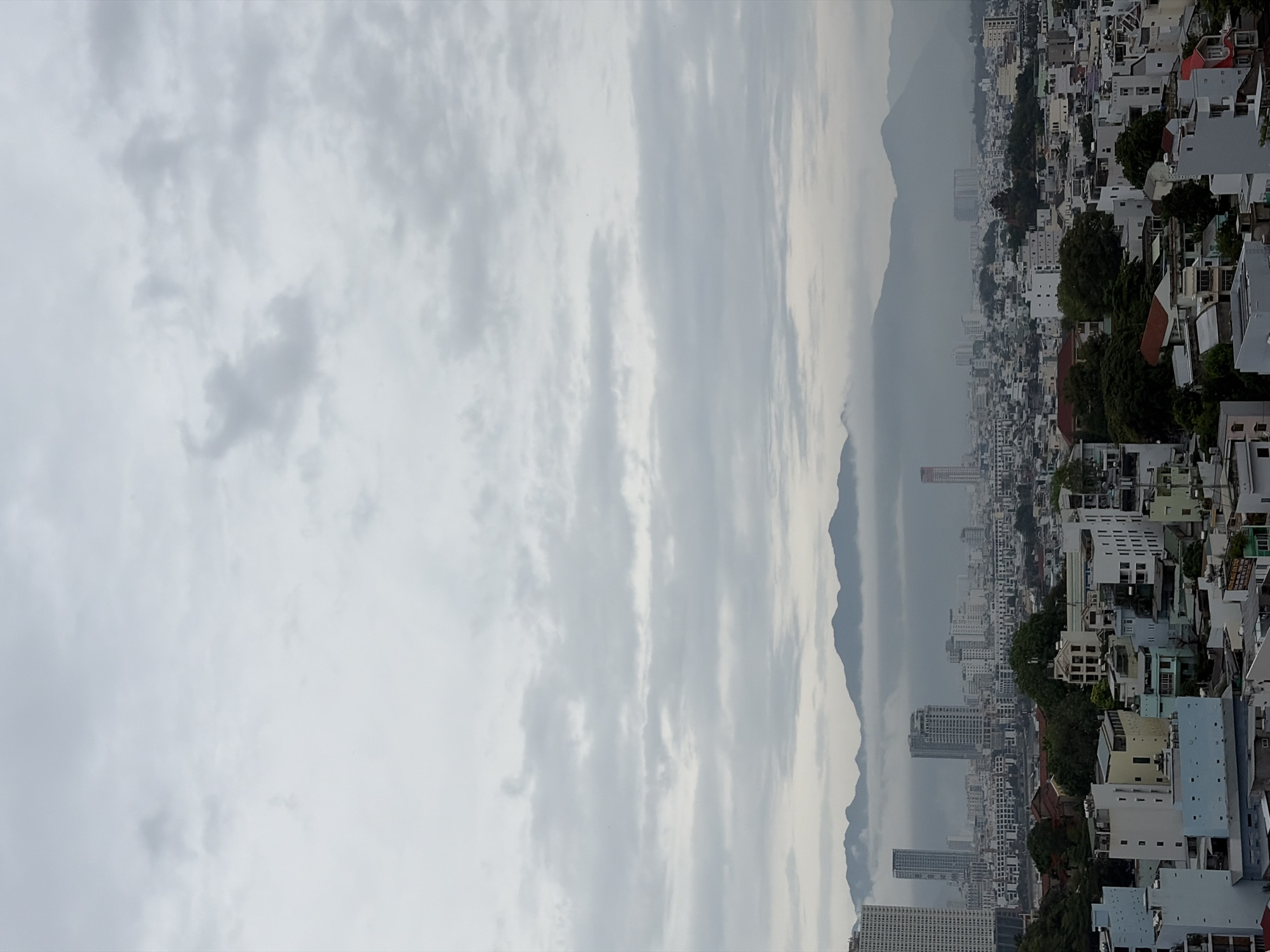 The image displays a panoramic view of an urban landscape, identified as Bac Nha Trang Ward, Vietnam, set against a backdrop of mountains under an overcast sky.
The upper portion of the frame is dominated by a dense, light grey cloud cover, indicating an overcast or heavily cloudy day. Below the sky, a range of mountains stretches horizontally across the mid-ground. These mountains appear somewhat hazy and subdued, blending into the atmospheric conditions.
The lower two-thirds of the image showcase the city, characterized by a high density of buildings. A mix of architectural styles is visible, including numerous low-rise residential structures with varied roof colors (predominantly white, grey, and some reddish-brown), alongside several taller, modern high-rise buildings that punctuate the skyline. Green foliage and trees are interspersed among the urban development, adding touches of nature to the concrete environment. The perspective suggests the photograph was taken from an elevated position, offering a sweeping view of the city and its surrounding natural features.
No specific human activity is visible, making the scene a static urban and natural landscape. The weather is distinctly overcast, contributing to a muted and slightly somber or tranquil mood. There is no discernible text within the image.