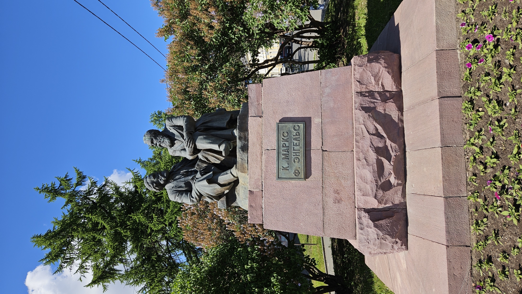 A bronze statue of two bearded men sitting side-by-side on a stone bench atop a large, multi-tiered stone monument is depicted outdoors. The men are dressed in formal attire, with one wearing a suit and the other a suit with a vest. Their faces are detailed, showing expressions of contemplation.
A plaque on the front of the monument reads "К. МАРКС Ф. ЭНГЕЛЬС" in embossed Cyrillic letters.
The monument is situated in a park-like setting with lush green trees and bushes surrounding it. A tall evergreen tree is visible to the left, and flowering bushes with small clusters of light-colored flowers are present behind the monument. The ground in front of the monument is a flowerbed with small green plants and a few scattered pink and purple flowers.
The sky above is a clear, bright blue with a few wispy white clouds. Electrical wires are visible against the sky in the upper right portion of the image. The overall scene is well-lit, suggesting a sunny day. The background shows glimpses of a street with buildings and vehicles, indicating an urban or suburban environment.