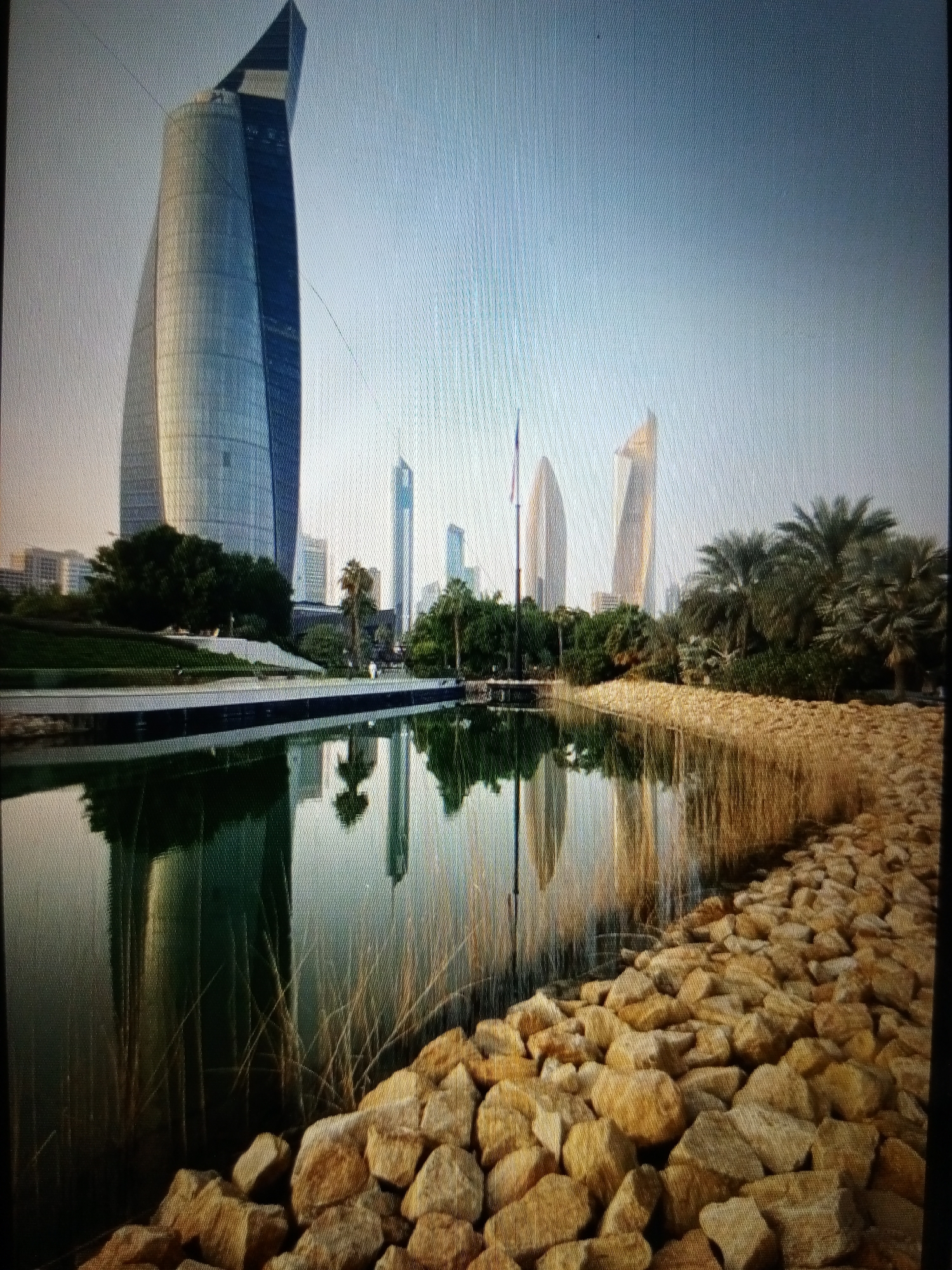The image displays a modern city skyline with several tall buildings, one of which is a distinctive glass skyscraper with a sharply angled top. In the foreground, a tranquil body of water reflects the sky and the surrounding architecture. The water's edge is lined with large, rough-hewn stones and tall, dry grasses. Beyond the water, a manicured green lawn slopes upwards towards the base of the buildings. Palm trees and other lush vegetation are interspersed throughout the landscape, creating a contrast between the natural elements and the urban structures. The sky is a pale, clear blue, suggesting a bright, possibly early morning or late afternoon, with a soft light illuminating the scene. There are no people visible in the image. The overall impression is one of serene modernity, with a blend of architectural grandeur and natural beauty. The image is not located in Golbembal, Nigeria.