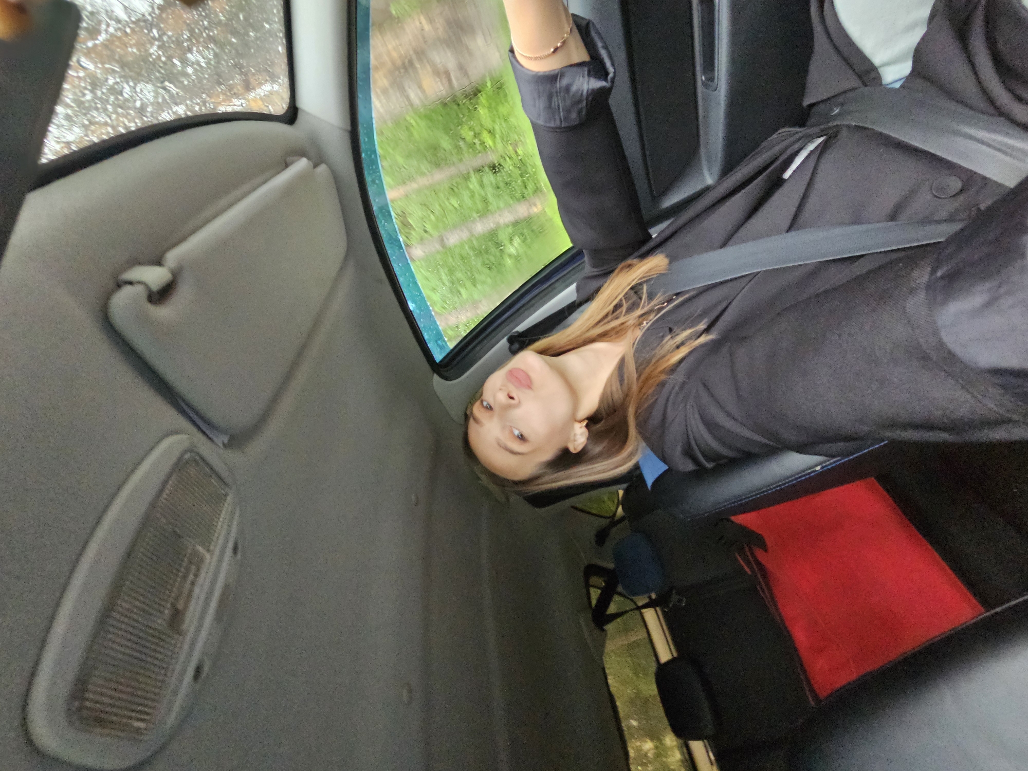 A young woman with long brown hair is seated in a car wearing a black jacket and a seatbelt. The car's interior is visible, including the sun visor, dome light, and window with a view of greenery outside. The location is the city of Chaikovsky, Russia.