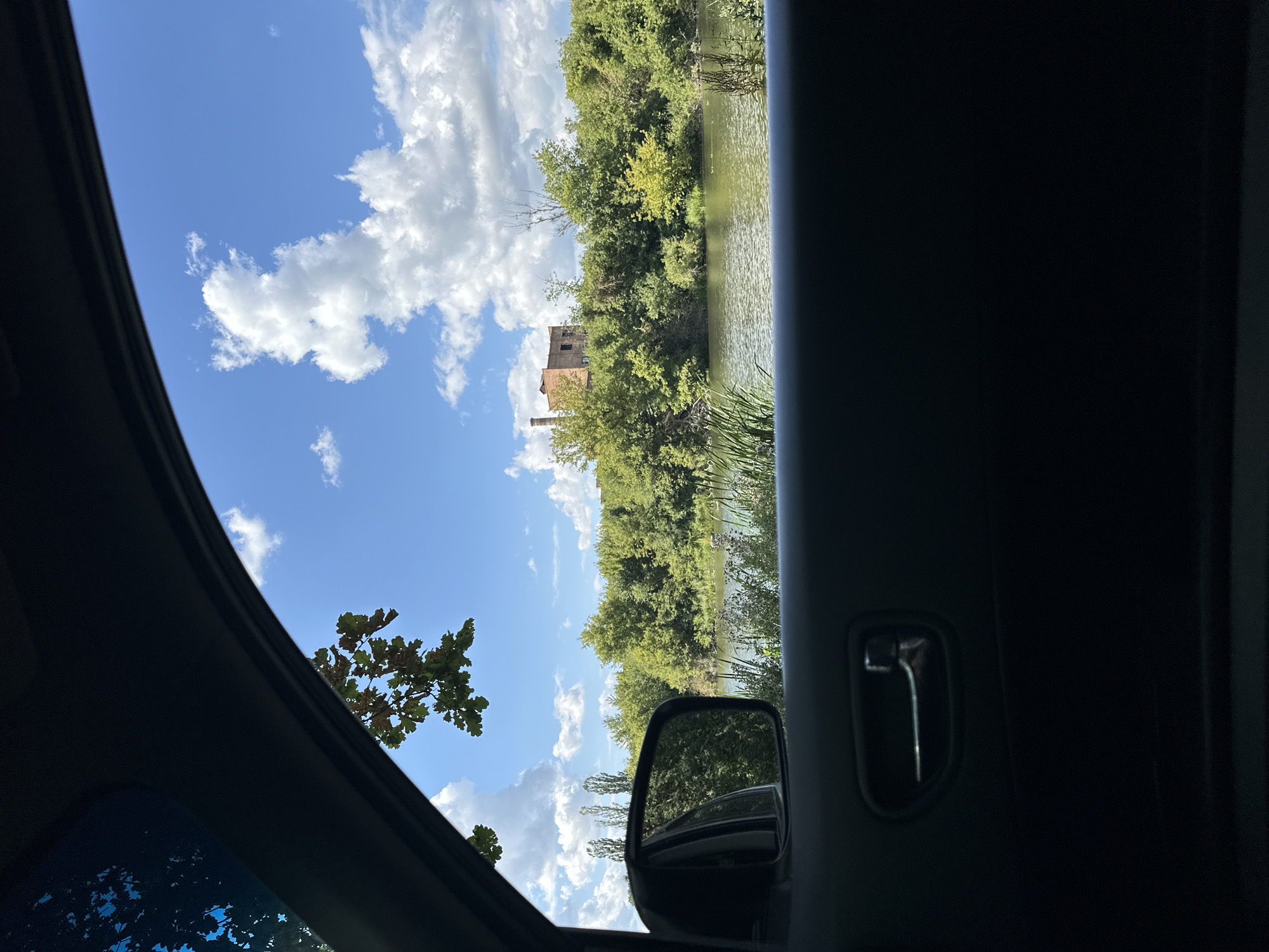 The image captures a view from inside a vehicle looking out through the side window. In the foreground, the dark interior of the car's door panel is visible, including a silver door handle. The window frames a scenic landscape. A body of water, likely a river, occupies the middle ground, with dense green foliage lining its banks. Beyond the trees, a dilapidated brick building with a prominent chimney stands on a slight rise, suggesting an abandoned industrial site or old structure. The sky is a vibrant blue, dotted with fluffy white clouds. A portion of a tree branch with green leaves is visible in the upper left corner, overhanging the car's interior. The side mirror of the car is also visible, reflecting the green trees and water. The scene depicts a rural or semi-rural setting, possibly on the outskirts of Shatava, Ukraine, given the architectural style of the building and the surrounding vegetation.