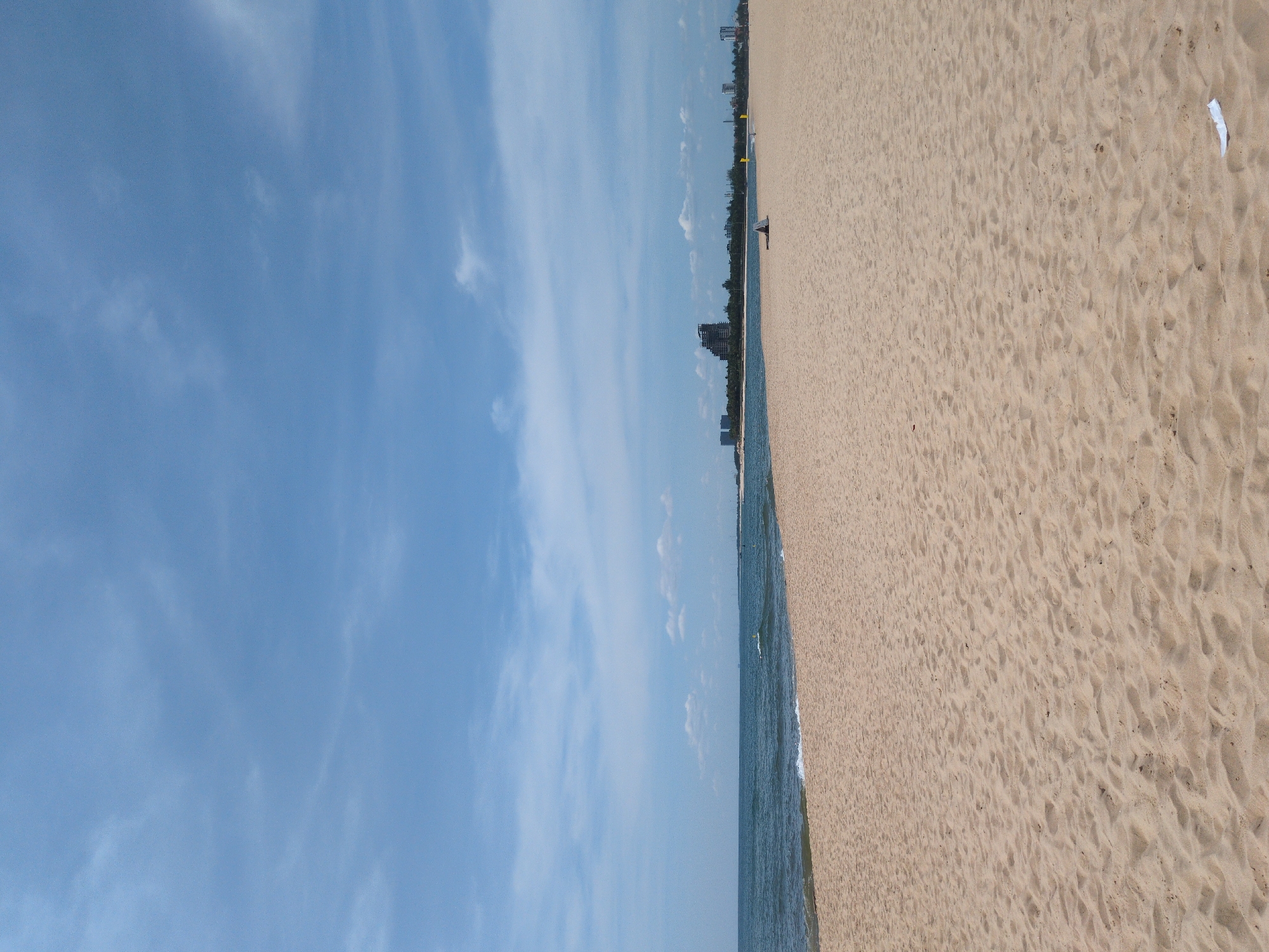 The image depicts a wide, sandy beach in Đồng Hới Ward, Vietnam, under a partly cloudy blue sky. The foreground is dominated by the textured sand of the beach, with subtle patterns from the tide or wind. In the middle ground, the calm, blue sea gently laps against the shore, with small waves breaking. Along the distant horizon, a line of trees separates the sea from a developed area with several buildings, including a large, unfinished-looking structure. A few individuals are visible on the beach: one person is seated under a small shelter, and another is further away, appearing to be walking or standing. A small white piece of debris is visible in the lower right corner of the sand. The overall scene conveys a sense of open space and a tranquil coastal environment.