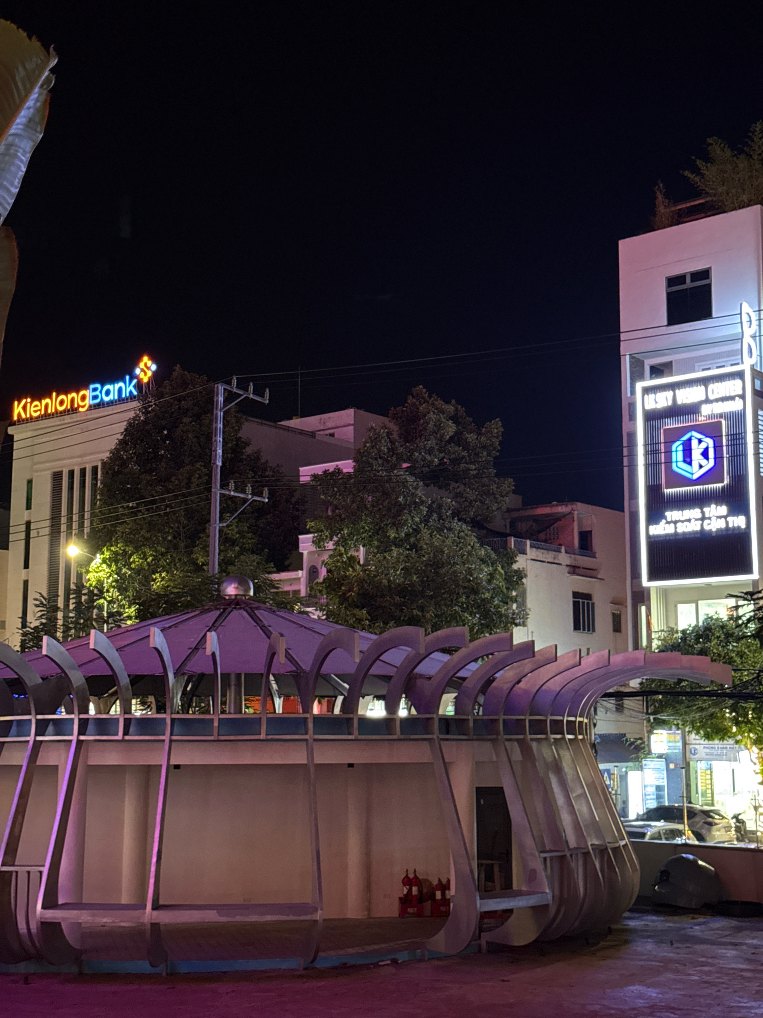 The image depicts a nighttime scene in the city of Nha Trang, Vietnam. In the foreground, a modern, open-air structure with a curved metal roof is visible. This structure appears to be a bus stop or waiting area, with benches and a light purple illumination. To the left, two red fire extinguishers are mounted on a white wall.
In the mid-ground, a large building with the neon sign "KienlongBank" is prominently displayed. The sign features orange and blue lettering with a dollar sign symbol. Across the street, another building displays a vertical neon sign that reads "LE SKY VISION CENTER" in white lettering, with a blue and white logo of a stylized "K" inside a square. Below this, text in Vietnamese, likely indicating a center name, is visible.
The background shows more buildings and trees, partially obscured by the night sky. Streetlights and other sources of artificial light illuminate the scene, creating a vibrant urban atmosphere. The overall impression is of a well-lit, modern commercial area at night.