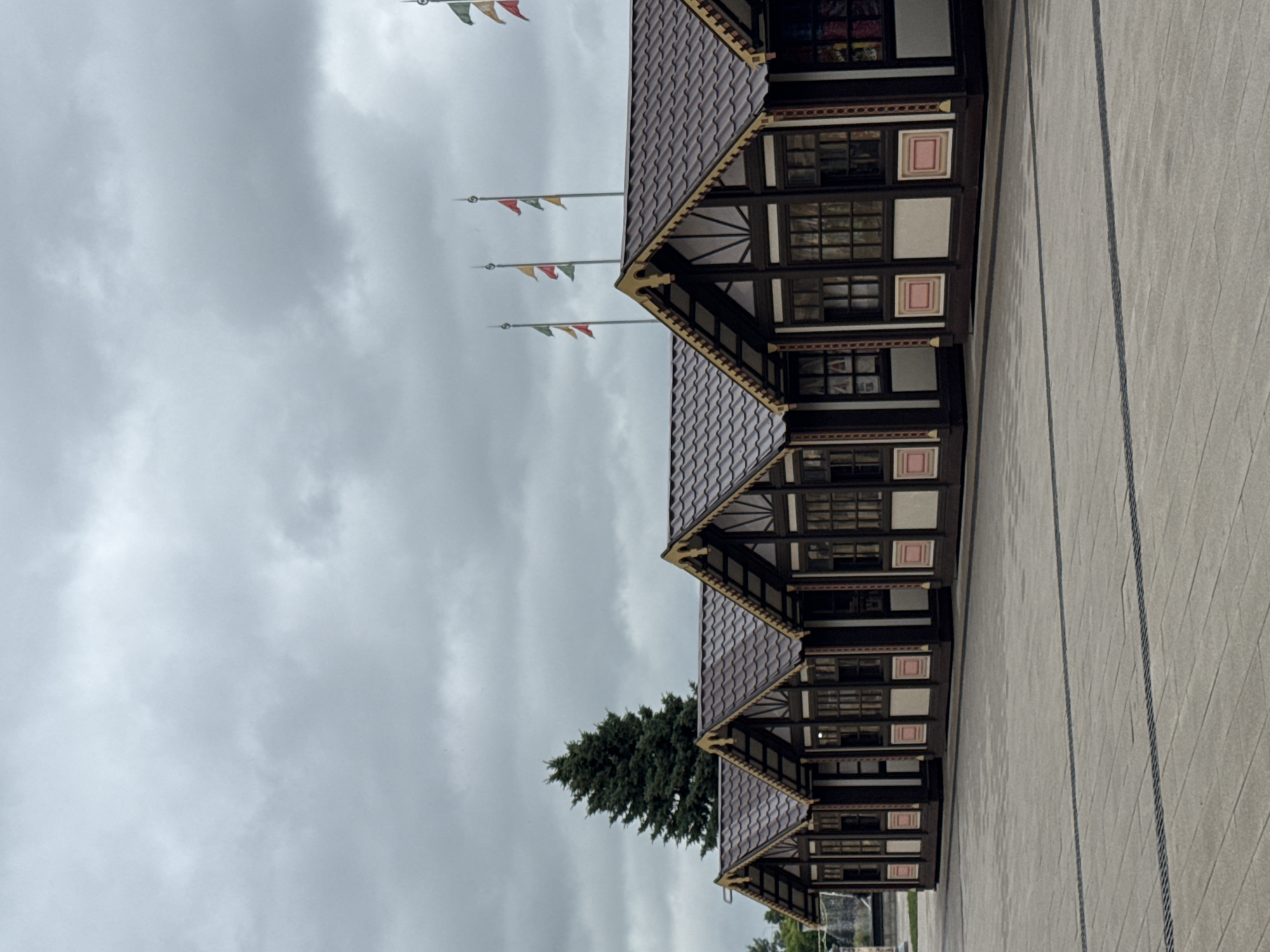 The image shows a row of small, dark brown market stalls with decorative trim and brown tiled roofs. The stalls have large windows with multiple panes. In the background, there is a large, dark green evergreen tree to the left, and several flagpoles with colorful flags flying from them on the right. The sky is overcast and gray. The ground in front of the stalls is a paved plaza with a repeating pattern of square stones and dark lines. The scene appears to be an outdoor market or fairground. The image does not provide enough visual information to determine the specific location in Nizhny Novgorod, Russia.