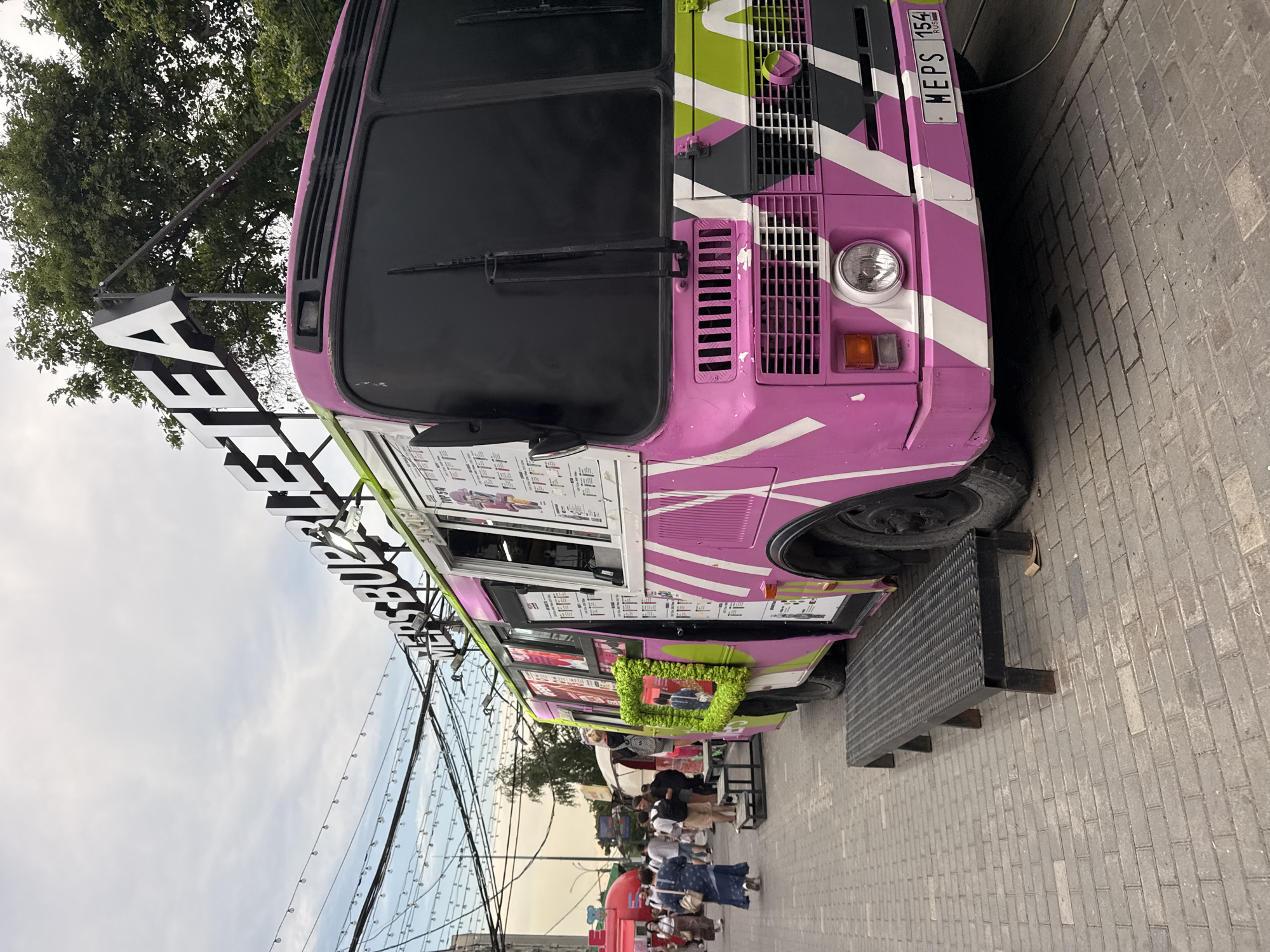 The image depicts a vibrant pink food truck, designed to resemble a bus, parked on a cobblestone street. The truck features bold white and lime green diagonal stripes. A prominent white sign above the bus reads "MER'S BUBBLE TEA." The license plate on the front of the truck displays "MEPS 154 RUS." In the background, there are other food vendors, including a red stall, and a crowd of people walking along the street. Overhead, string lights are visible, suggesting an outdoor market or festival atmosphere. The overall scene is bustling and colorful, indicative of a lively urban setting. Based on the license plate and the architectural style of the surrounding stalls, this scene is likely located in Novosibirsk, Russia.