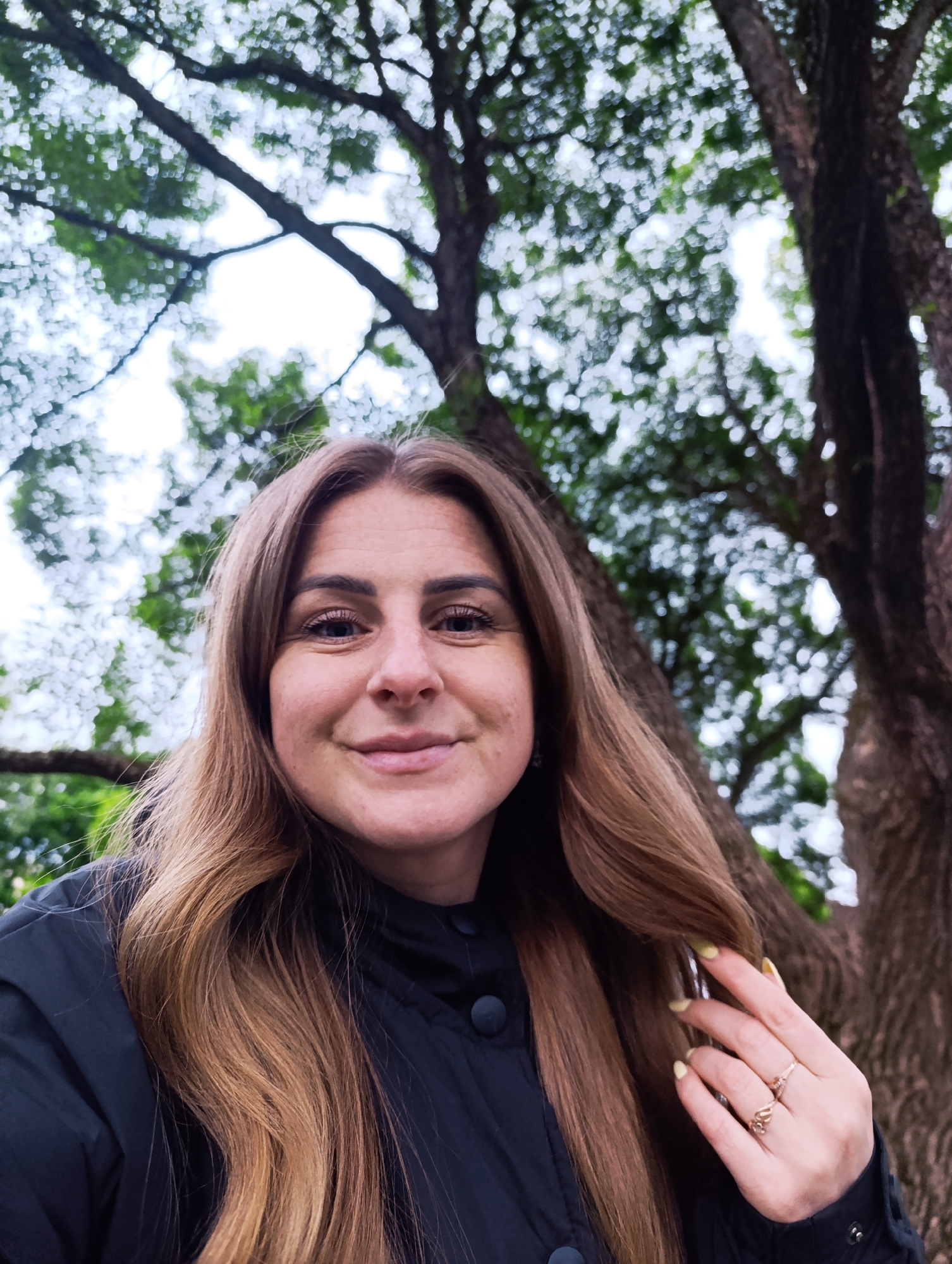 A woman stands beneath the canopy of a large tree. The tree has a thick trunk and expansive branches with green foliage. She is fair-skinned with brown hair, wearing a dark jacket and two rings on her right hand, her fingernails painted a light color. The location appears to be a park or natural area, and the sky is overcast.