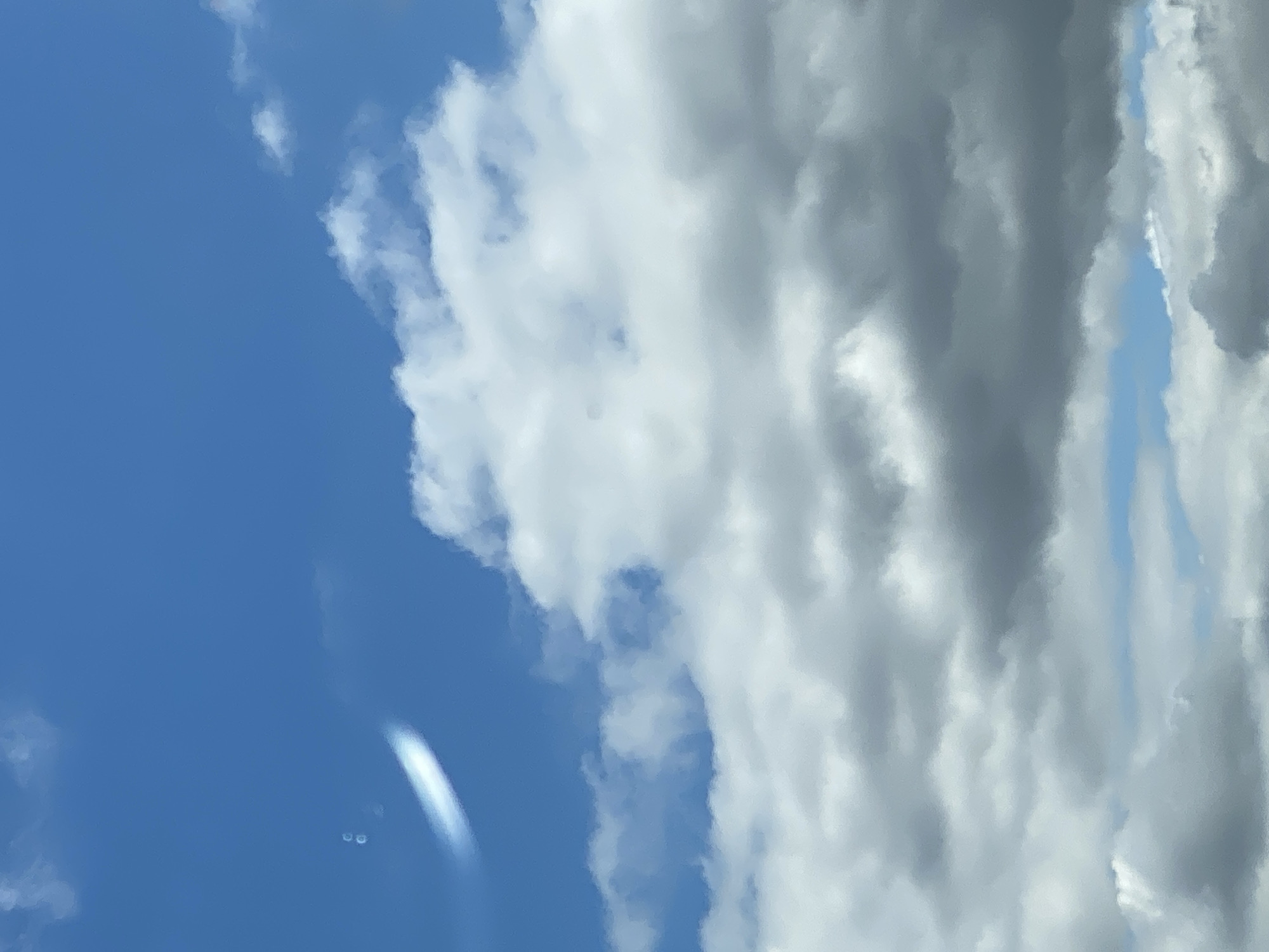 The image shows a blue sky with scattered white and grey clouds. There are two blurry white oval shapes in the upper left quadrant of the image, one larger than the other. It is unclear if these are lens flares or some atmospheric phenomenon. The lower portion of the image is dominated by large, puffy white clouds, some of which appear to be casting shadows, creating areas of grey. The scene appears to be an outdoor shot, likely taken during the daytime, with no people or man-made objects clearly visible. The image is too generic to determine a specific location within Nossen, Germany.