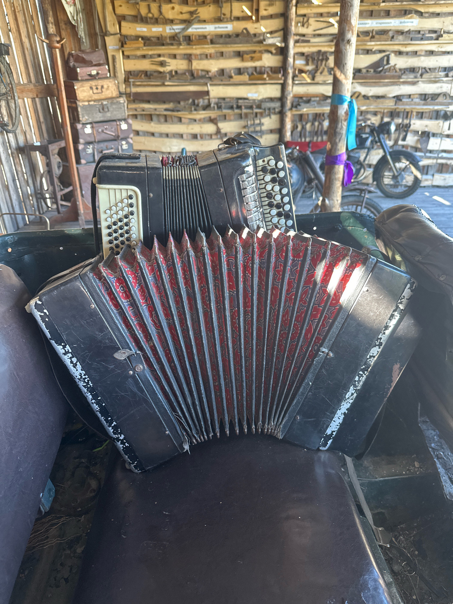 An accordion rests on a dark, padded surface, possibly a seat, within what appears to be a workshop or storage area. The accordion is black with a red and patterned bellows. Behind the accordion, the wall is lined with shelves holding various tools and objects, some of which appear to be antique. A wooden pole with two colored ribbons (blue and purple) is visible in the center of the background. To the right of the pole, a motorcycle is parked. The overall impression is one of a cluttered, rustic setting, possibly in the city of Kargat, Russia. The lighting suggests it is daytime, with sunlight casting shadows.