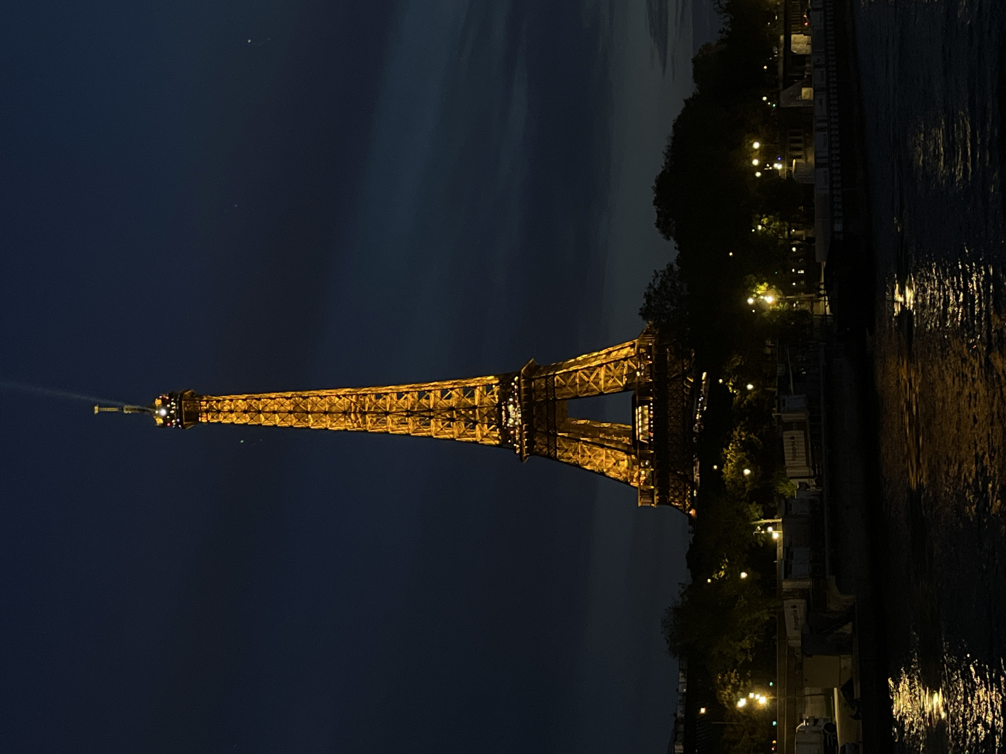 This media file presents a striking vertical composition of the illuminated Eiffel Tower at night, observed from across a body of water, likely the Seine River in Paris, France. The iconic landmark stands prominently in the center-left, glowing with a warm, golden light that highlights its intricate lattice structure. A powerful beam of light emanates from the very top of the tower, extending upwards into the dark night sky.
The sky is a deep, dark blue, transitioning to lighter hues towards the horizon, suggesting either a very late twilight or early dawn, though primarily appearing as night. Faint cloud formations are visible in the upper sections of the sky.
To the right of the Eiffel Tower, the surface of the river is visible, reflecting the golden lights of the tower and other scattered lights along the riverbanks, creating shimmering, elongated patterns on the water. The riverbank itself, on the side opposite the viewer, is lined with dark silhouettes of trees and indistinct buildings, dotted with numerous bright, yellowish streetlights or lights from structures. These lights cast their own reflections onto the water.
The overall scene captures a classic and romantic nighttime view of the Eiffel Tower in its Parisian setting. No people are visible in the image, and there is no discernible text. The image conveys a calm and atmospheric moment.