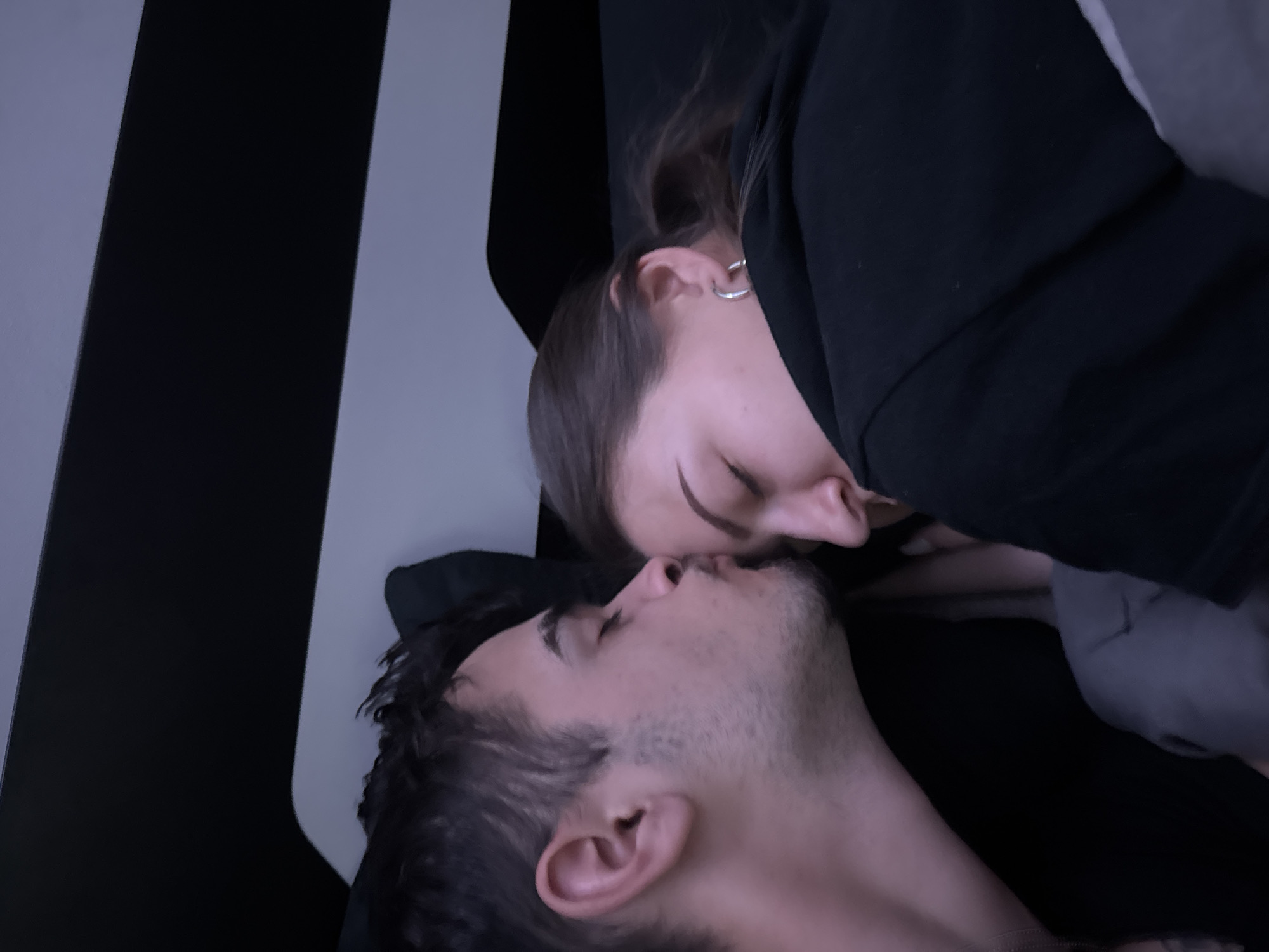 A couple is shown in close proximity. The woman is on top with her face close to the man's face. They appear to be kissing or about to kiss. The woman has an earring and is wearing a black sweatshirt. The man has short, dark hair and stubble. The background includes a black and white striped pattern. There is no discernible information that indicates the location is Sofia, Bulgaria.