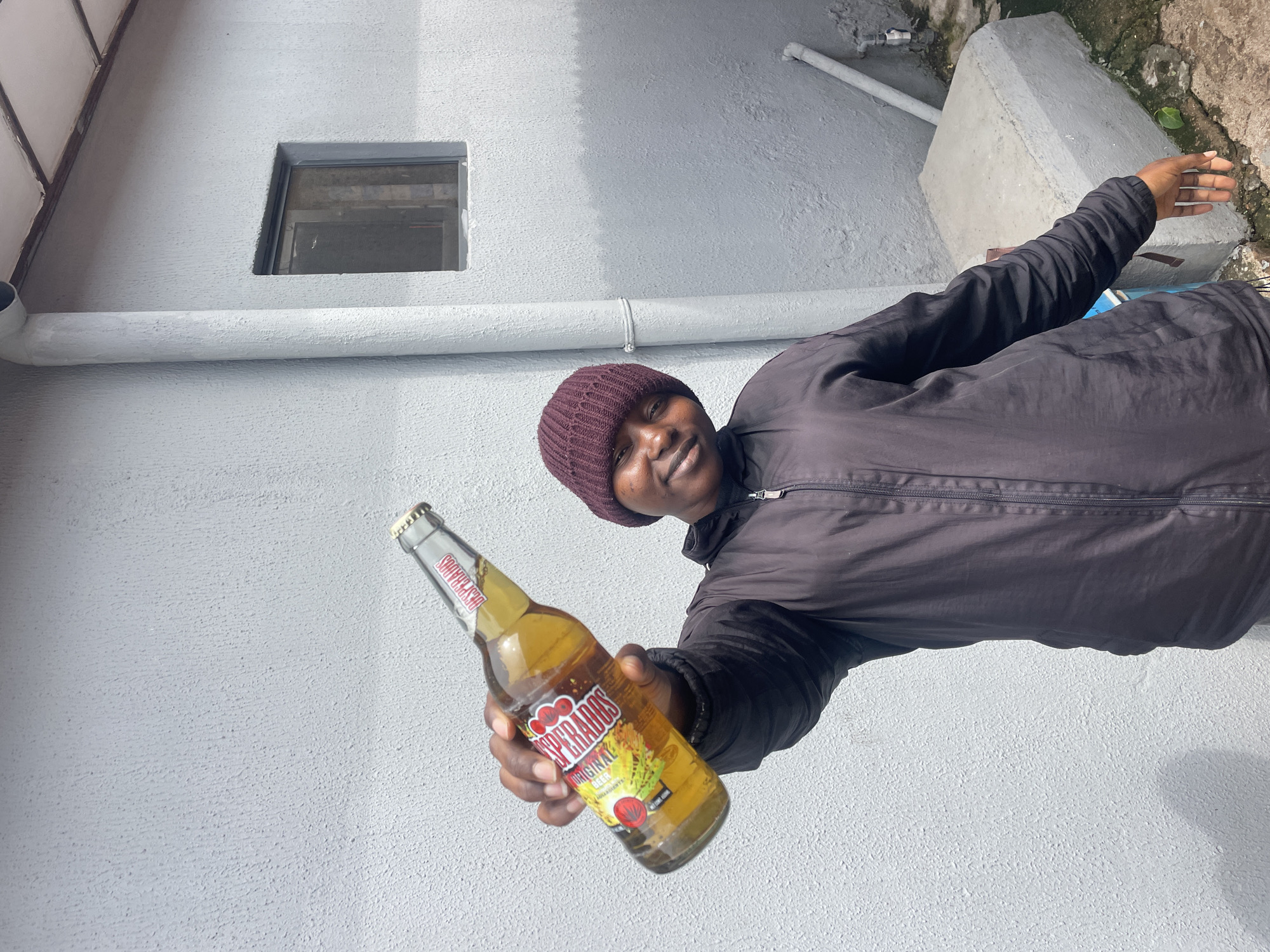 This daytime photograph, taken in Jos, Nigeria, features a person holding out a bottle of "Desperados Original Beer" towards the viewer. The individual has dark skin, a friendly smile or smirk, and is looking directly at the camera. They are wearing a dark maroon knitted beanie and a dark, possibly black or very dark brown, zip-up jacket.
The glass bottle of beer is clearly labeled "DESPERADOS ORIGINAL BEER" with smaller text indicating it is "BEER FLAVOURED WITH TEQUILA" and an "ALCOHOLIC DRINK." The label features colorful graphics, including red and yellow elements, possibly depicting agave or chili peppers.
The setting is outdoors, against a light gray or off-white textured wall, likely made of stucco or painted concrete. A white horizontal pipe runs along this wall. Above the pipe, a small, recessed rectangular window with a dark frame is visible. In the upper left, part of a multi-paneled structure, possibly a roof extension or awning, can be seen. To the right of the person, the ground consists of a concrete block structure and then transitions into a rougher area with dirt and minimal foliage, suggesting a functional or utility space beside a building.
The bright, even lighting indicates a clear, sunny day, contributing to the casual and inviting atmosphere conveyed by the person's expression and gesture.