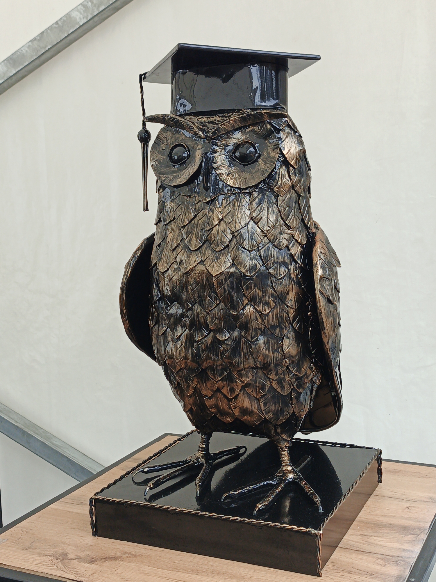 A metallic sculpture of an owl wearing a graduation cap stands on a wooden surface. The owl is depicted with intricate feather detailing and appears to be made of bronze or a similar metal. It has large, dark eyes and a black graduation cap with a tassel. The sculpture is positioned on a black, glossy base. The background consists of a plain white wall and a metal beam. This owl sculpture is located at the Zlatoust Museum of Local Lore, in Zlatoust, Russia.