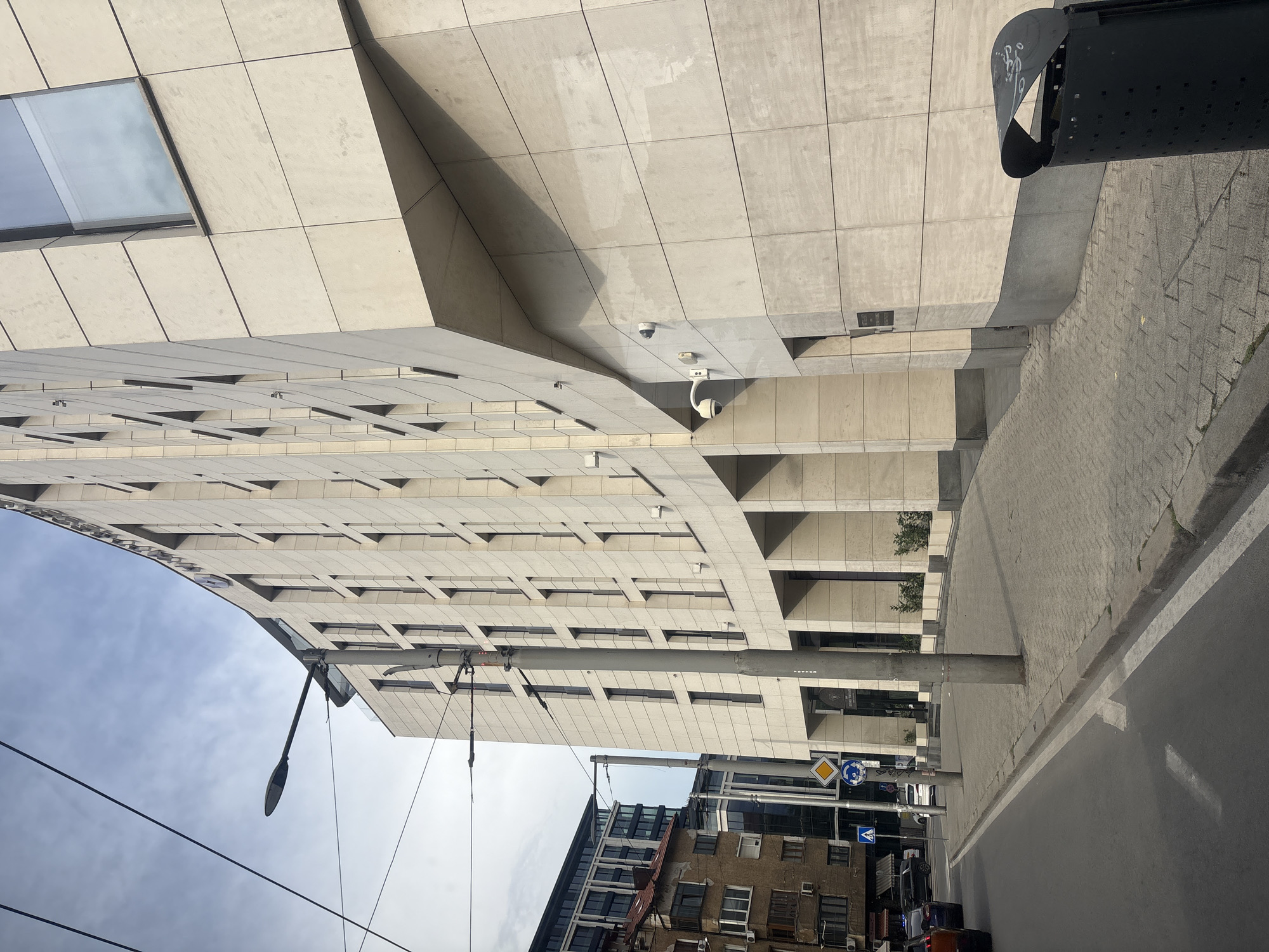 The image captures a street scene in Sofia, Bulgaria, with a focus on a modern, light-colored stone-clad building. The building has a curved facade with numerous vertical rectangular windows. Below the main structure, there are arched openings creating a colonnade effect. Two security cameras are visible on the exterior of this building. To the left of the modern building, an older, dilapidated building with visible signs of wear and tear is partially visible, featuring a mix of windows and balconies. A street lamp on a pole stands prominently in the foreground, with overhead wires crisscrossing the sky. The street itself is paved, with a sidewalk made of interlocking bricks. Traffic signs, including a yellow diamond-shaped sign with a black border and a blue circular sign with an arrow indicating a roundabout, are present. Several cars are on the road, and a black trash receptacle is positioned on the sidewalk. The sky is overcast with some blue patches. The scene depicts an urban environment with a juxtaposition of old and new architecture.