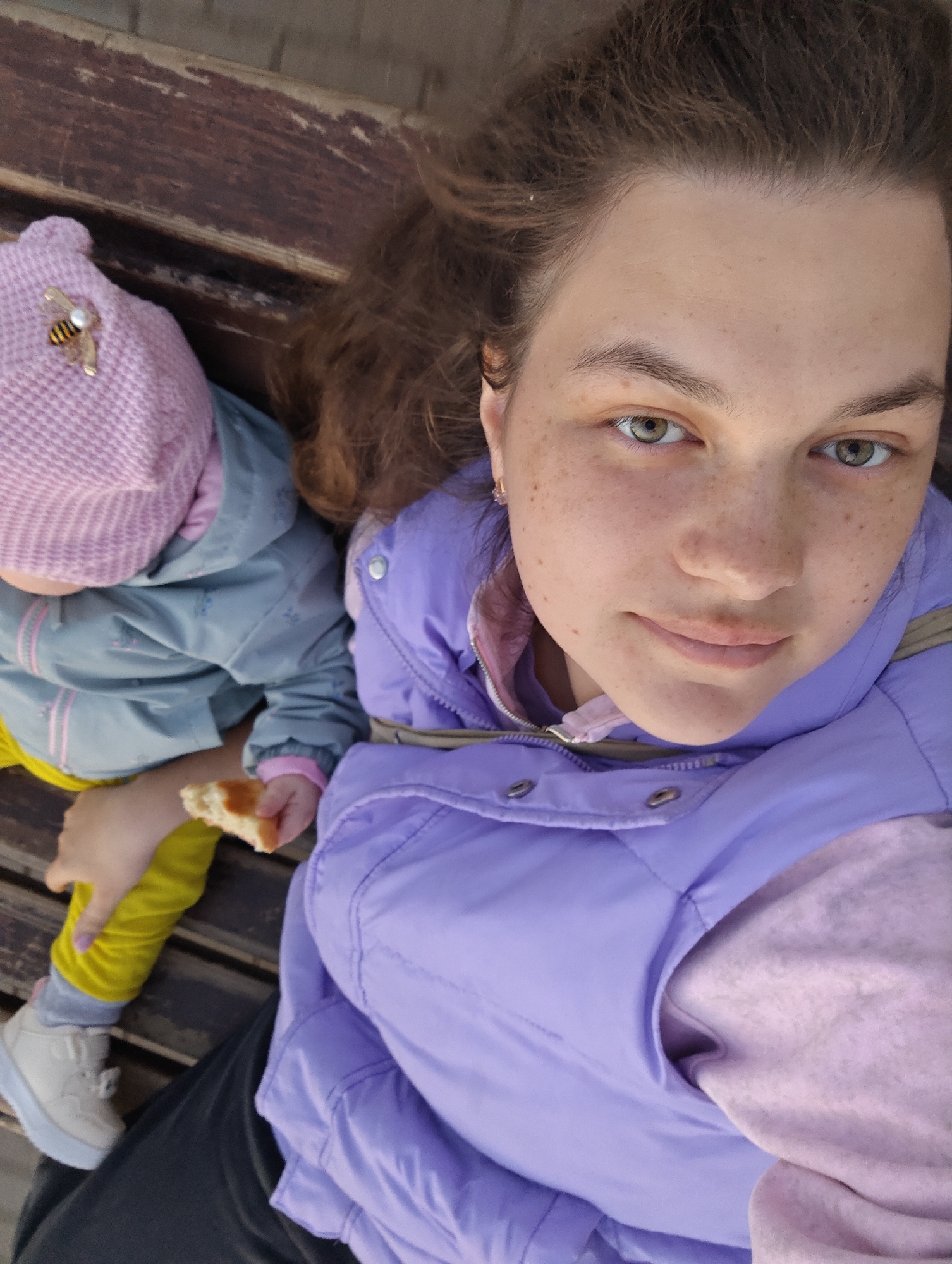 A medium-shot photograph shows a person and a child sitting on a wooden bench outdoors.
The person, likely a young adult woman, is on the right side of the image, wearing a light purple puffer vest over a similarly colored hoodie. She has brown hair and fair skin with freckles. Her face is directed towards the camera with a slight smile.
To the left, a toddler-aged child sits, wearing a pink knitted hat with a bee-shaped decoration, a gray jacket with pink accents, and yellow pants. The child is holding a piece of bread in their hand. A white shoe is visible on one of the child’s feet.
The background is a wooden bench with gaps between the slats. The lighting appears to be soft and natural.
The location is determined to be in the city of Chaikovsky, Russia. However, this is not evident from the visual information provided in the image alone. Additional contextual information not included in the image must have been used to make that determination.