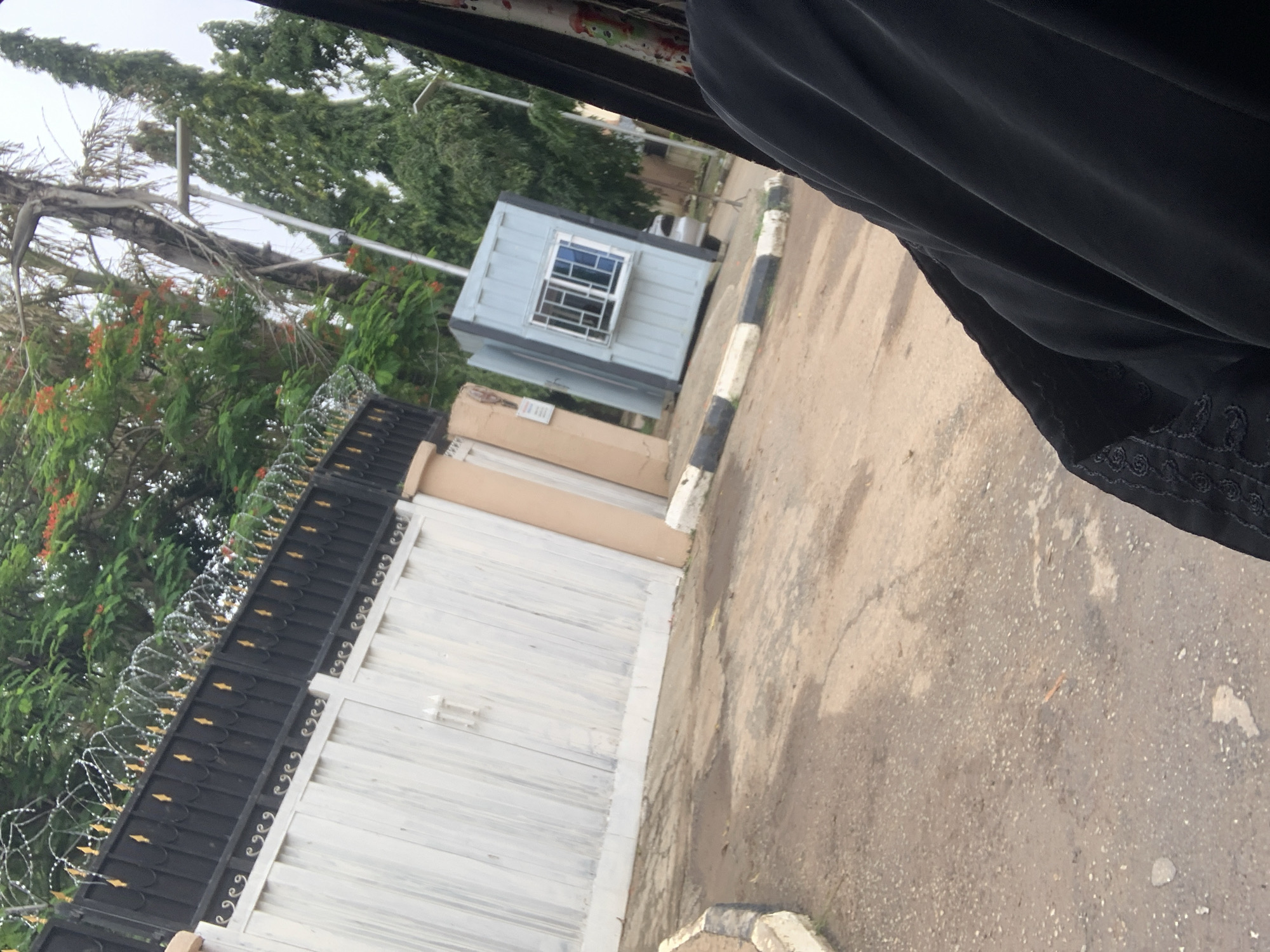 The image shows a partial view of an outdoor scene, likely in Abuja, Nigeria, as suggested by the context. The perspective is from inside a vehicle, with a black fabric partially obscuring the top and right side of the frame, suggesting the viewer is looking out a window.
The main subjects in view are a black metal gate with decorative gold elements and barbed wire on top, a large white gate with vertical wooden planks, a small light blue security booth with a barred window, and a backdrop of lush green trees, some with red flowers. The ground appears to be an asphalt road with some cracks and patches.
The security booth is positioned to the left of the road, near the gates. The gates seem to be for a property entrance. The presence of barbed wire on the black gate indicates a focus on security. The overall scene suggests a daytime setting, with overcast or diffused lighting, as there are no strong shadows.
No specific activity is clearly depicted, but the setting implies access to a property, possibly a residence or commercial establishment, with security personnel likely stationed in the booth. The details of the gates, the booth, and the vegetation are the most prominent visual elements. There is no discernible text in the image.